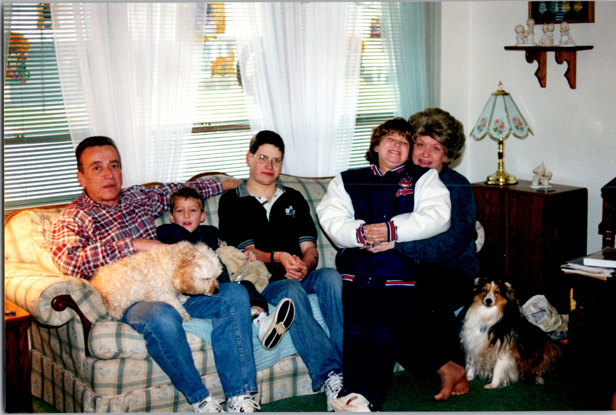 Multi-generational family on a plaid sofa in a living room; a beige fluffy dog rests on the man’s lap, with a dog on the floor, and two women with a teen nearby.