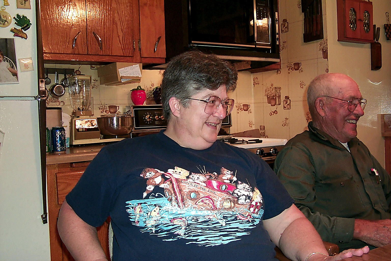 Two adults smiling and talking in a home kitchen; a woman in a navy graphic T‑shirt sits at the counter beside a man in a green shirt, with wooden cabinets and appliances in the background.
