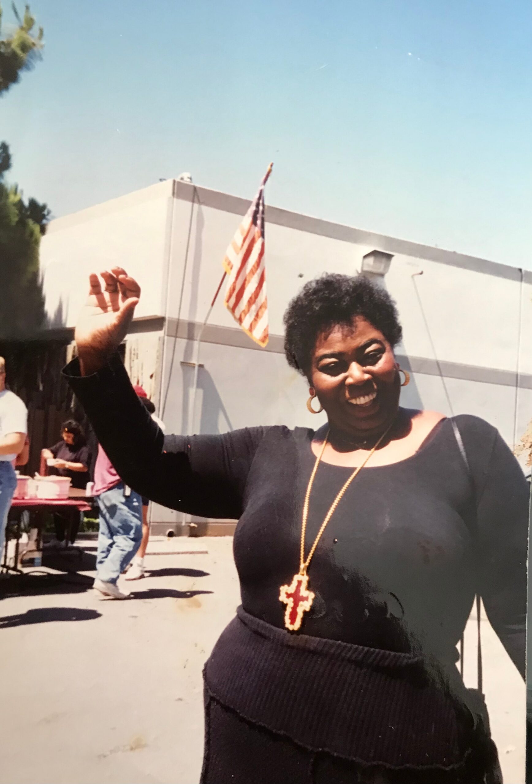 Smiling Black woman in a black top waves with a gold cross necklace, outdoors with an American flag in the background.