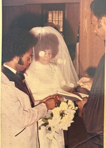 Bride in veil and white dress, groom in a suit, and officiant reading from a book during an indoor wedding ceremony.