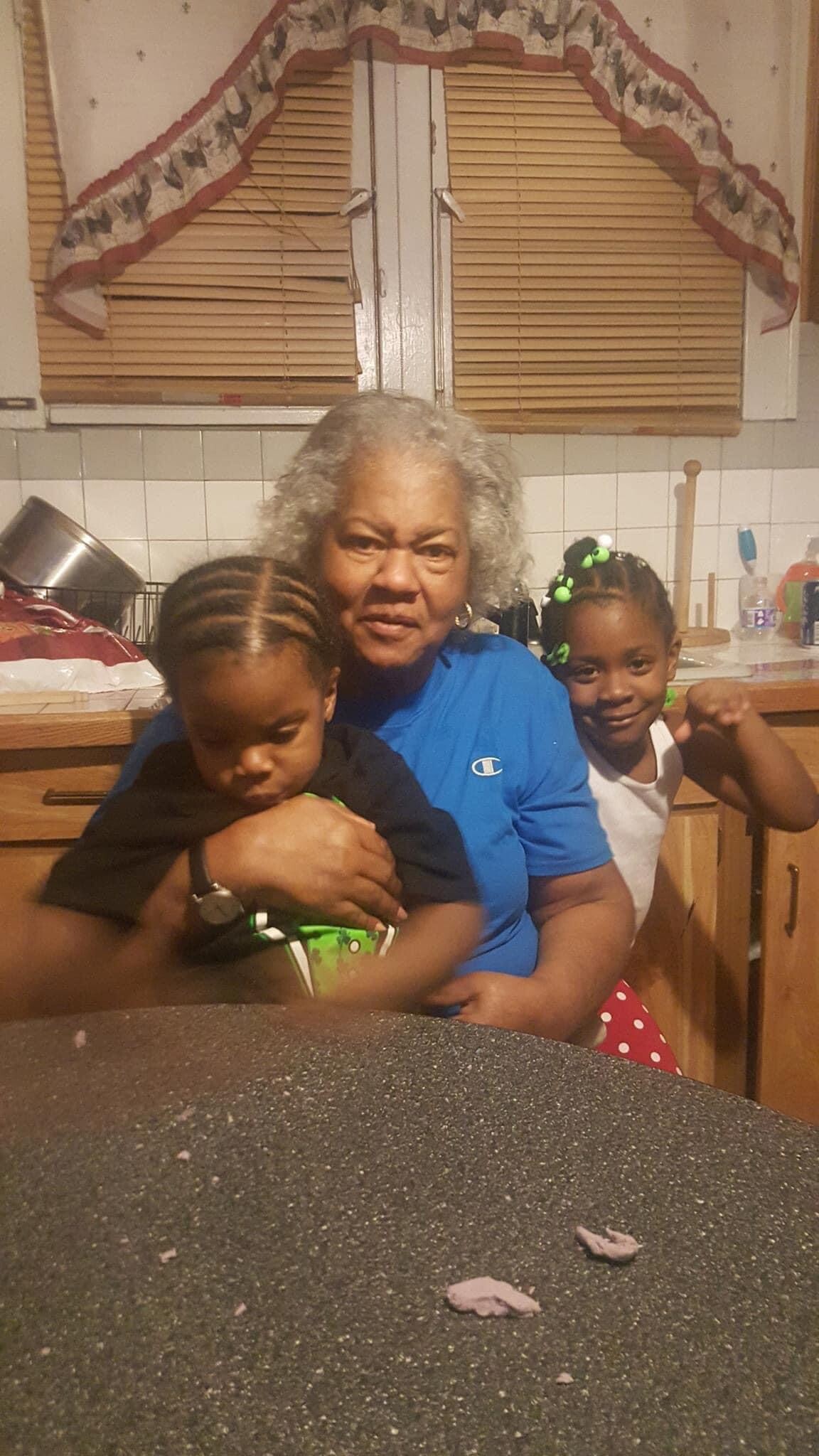 Grandmother in a blue shirt sits at a kitchen table with two children, one resting on her lap and the other smiling beside them.