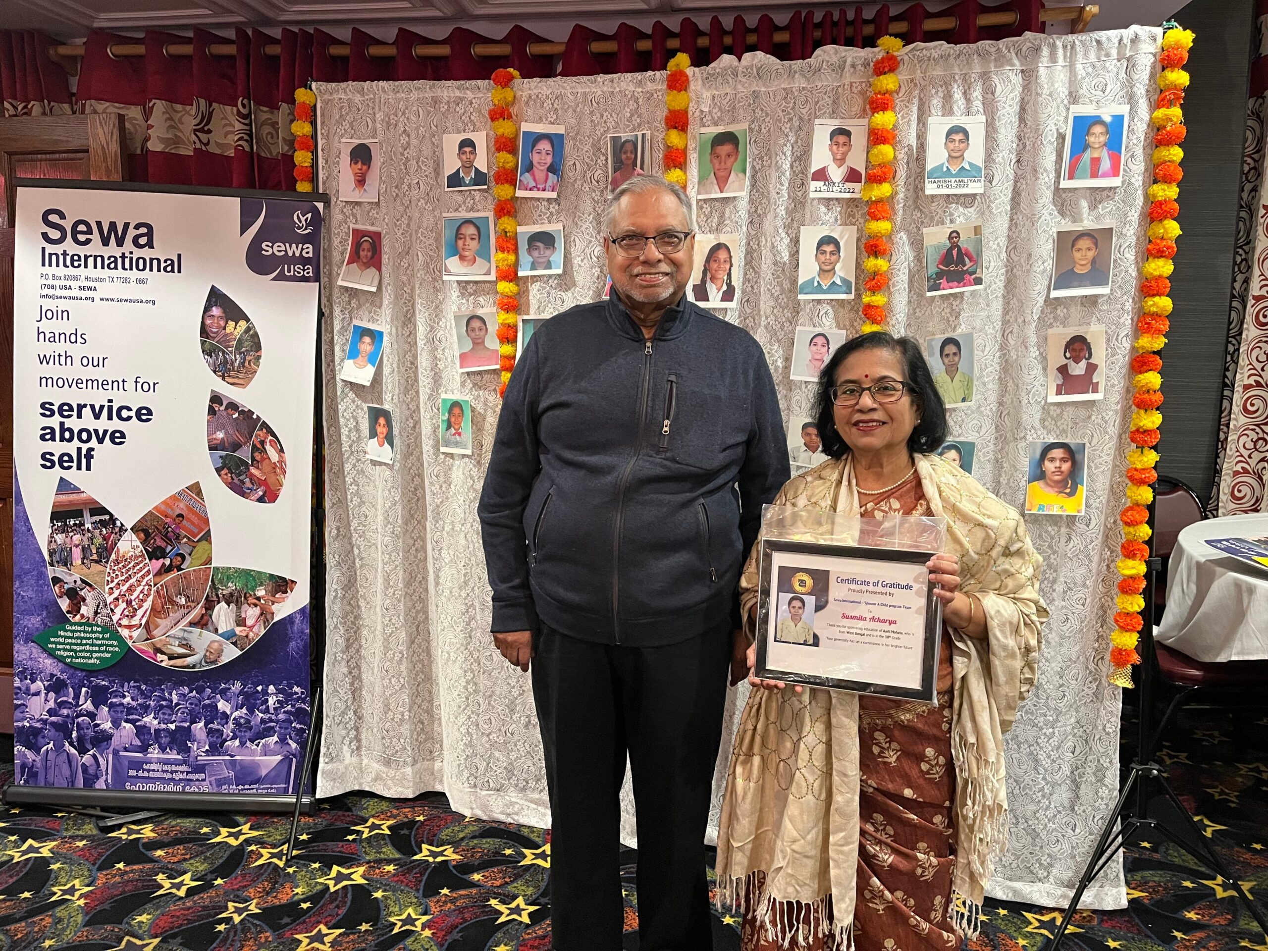 Man and woman pose in front of a photo-wall backdrop with marigold garlands at a charity event; banner reads Sewa International on the left.