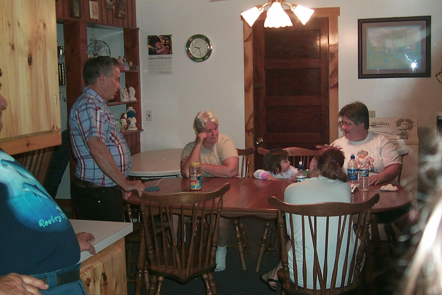 Family and friends gather around a wooden dining table in a kitchen, chatting and relaxing indoors.