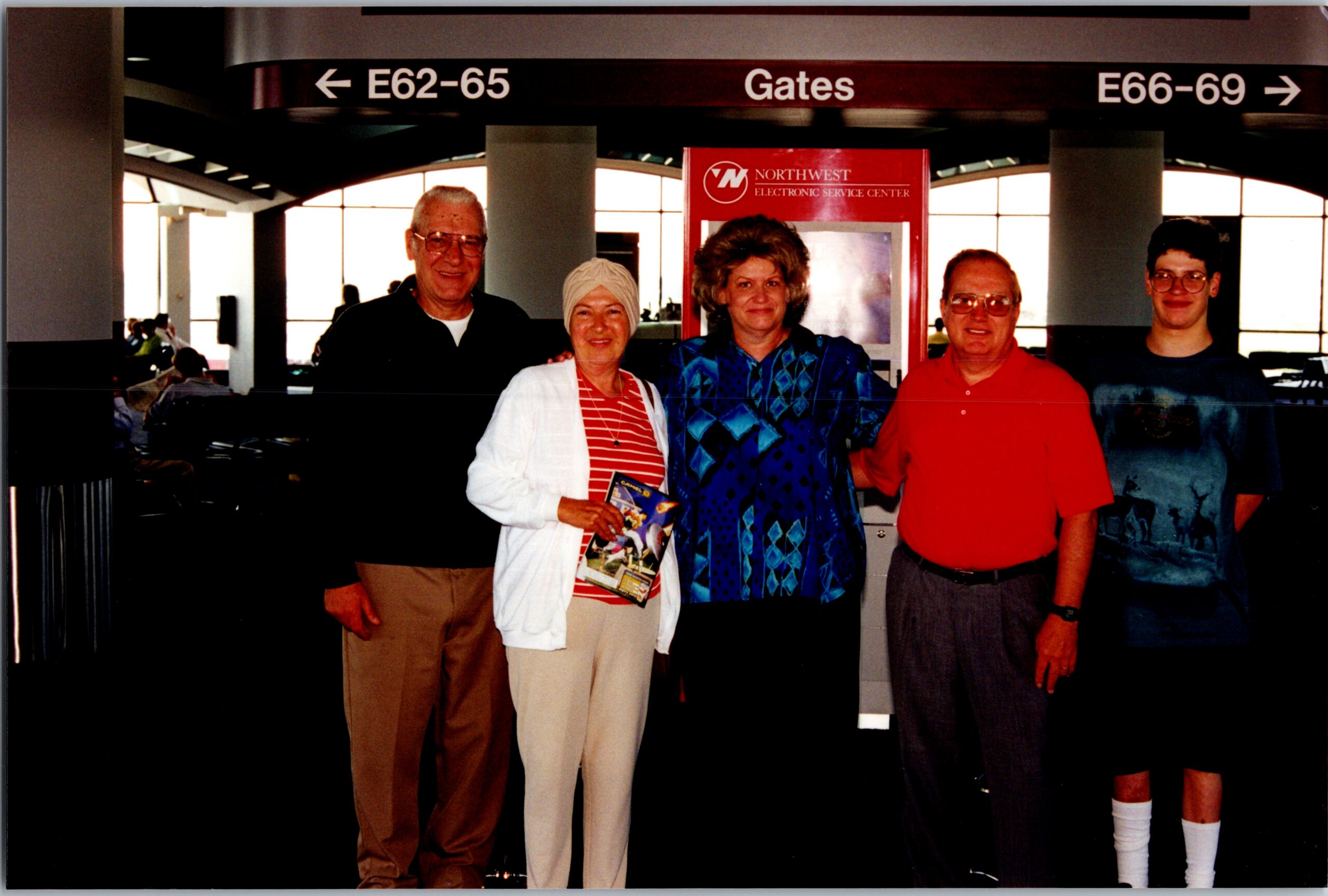 Five adults pose together in an airport terminal by a Gates sign and a red Northwest Electronics display behind them.