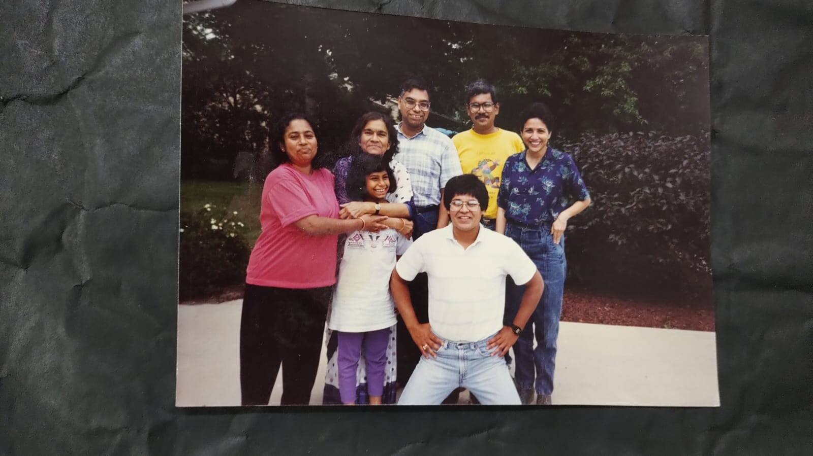 Seven people pose together outdoors, smiling, in a garden-like setting with bushes in the background.