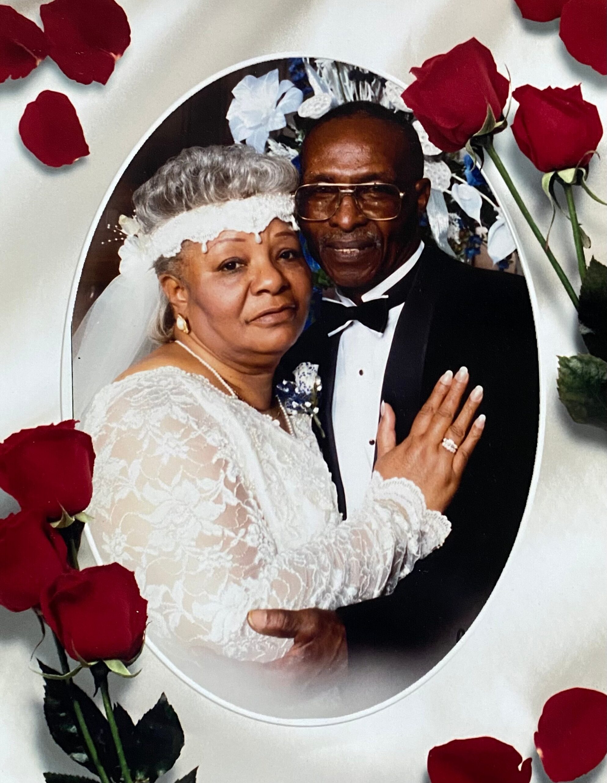 A smiling elderly couple posing in wedding attire inside an oval frame, surrounded by red rose petals.