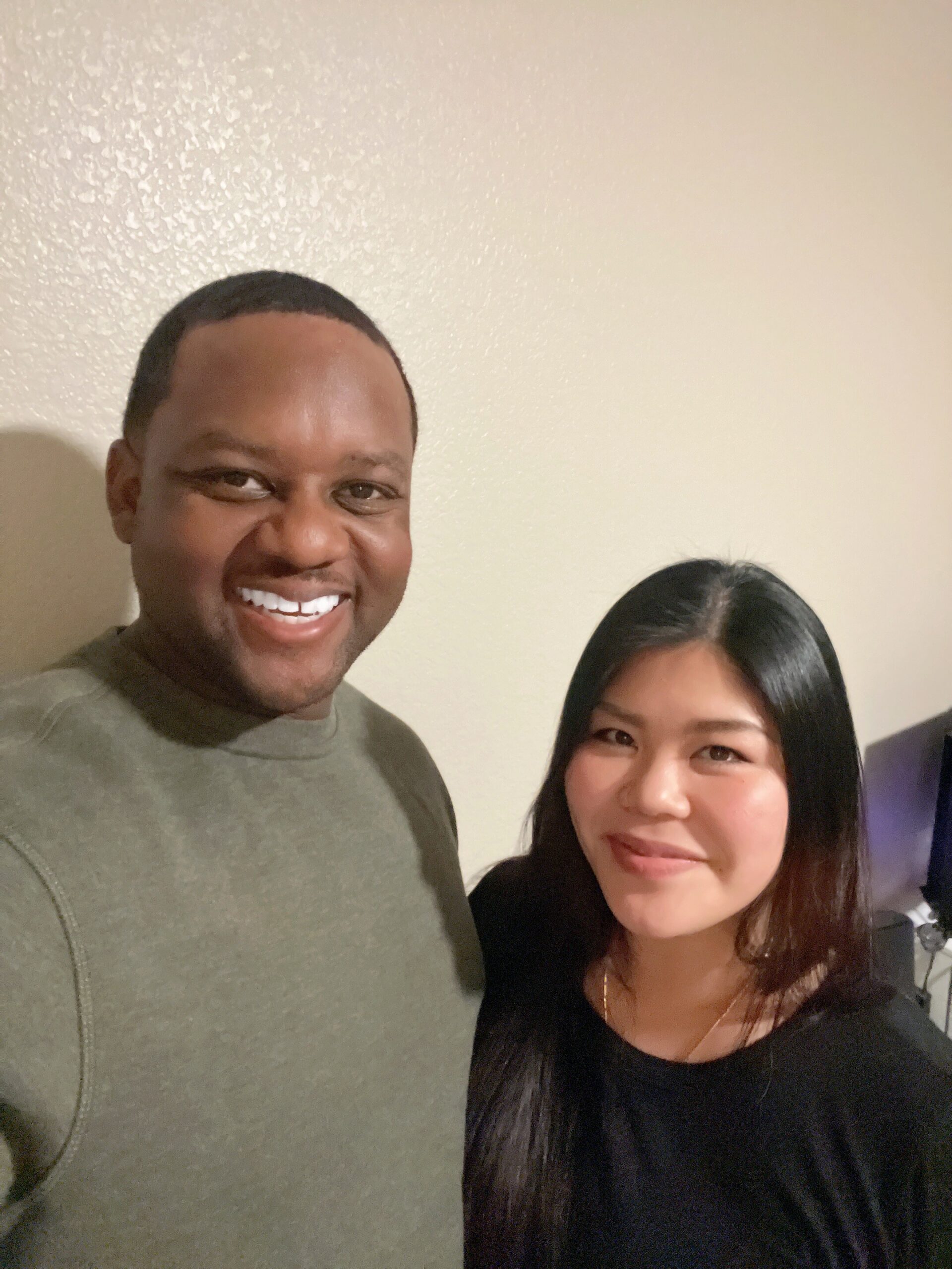 Smiling man and woman posing for a selfie indoors against a beige wall.