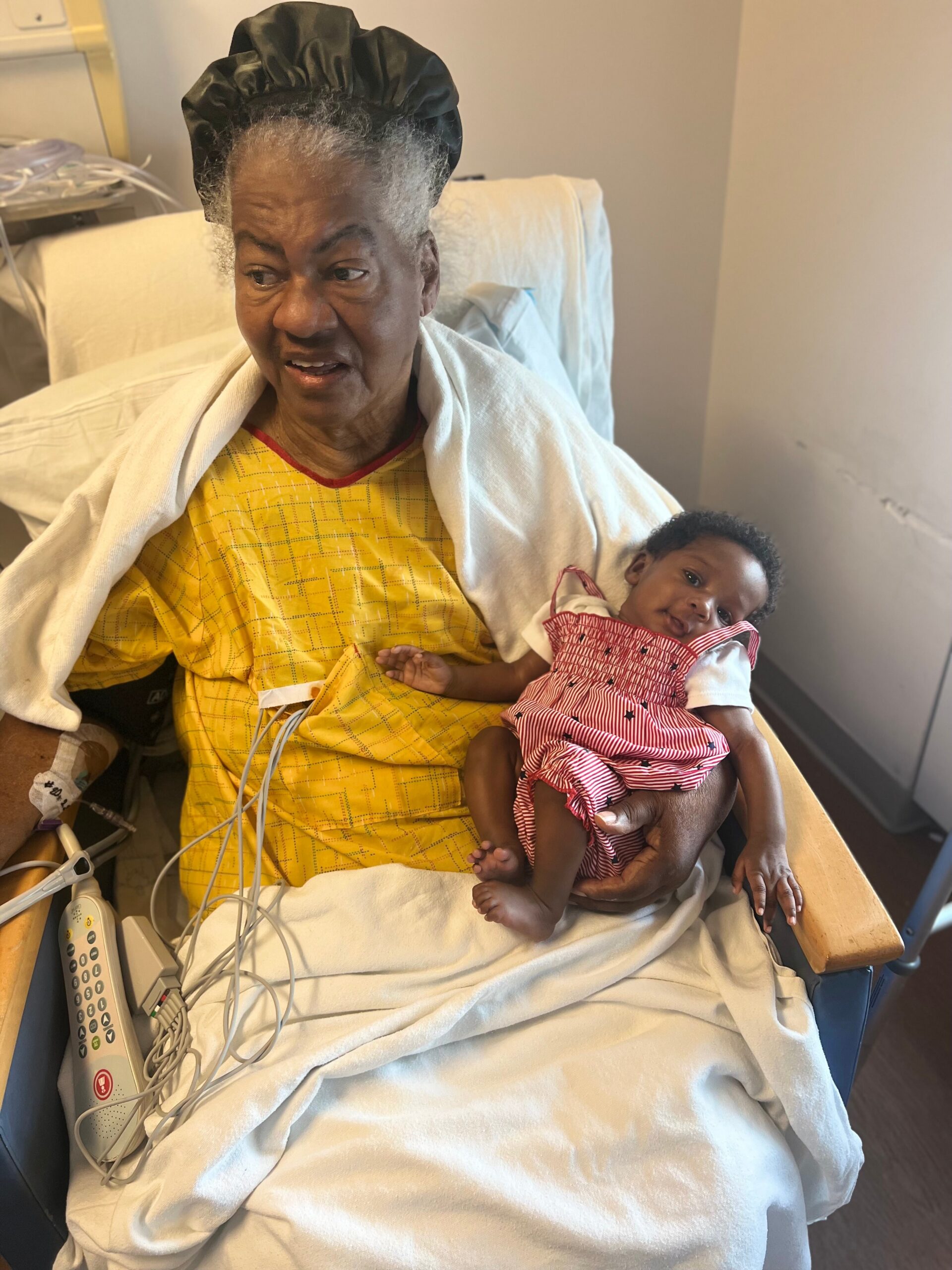 Older woman in a hospital bed wearing a yellow gown and black cap holds a baby dressed in a red-and-white striped outfit, with medical tubing nearby.