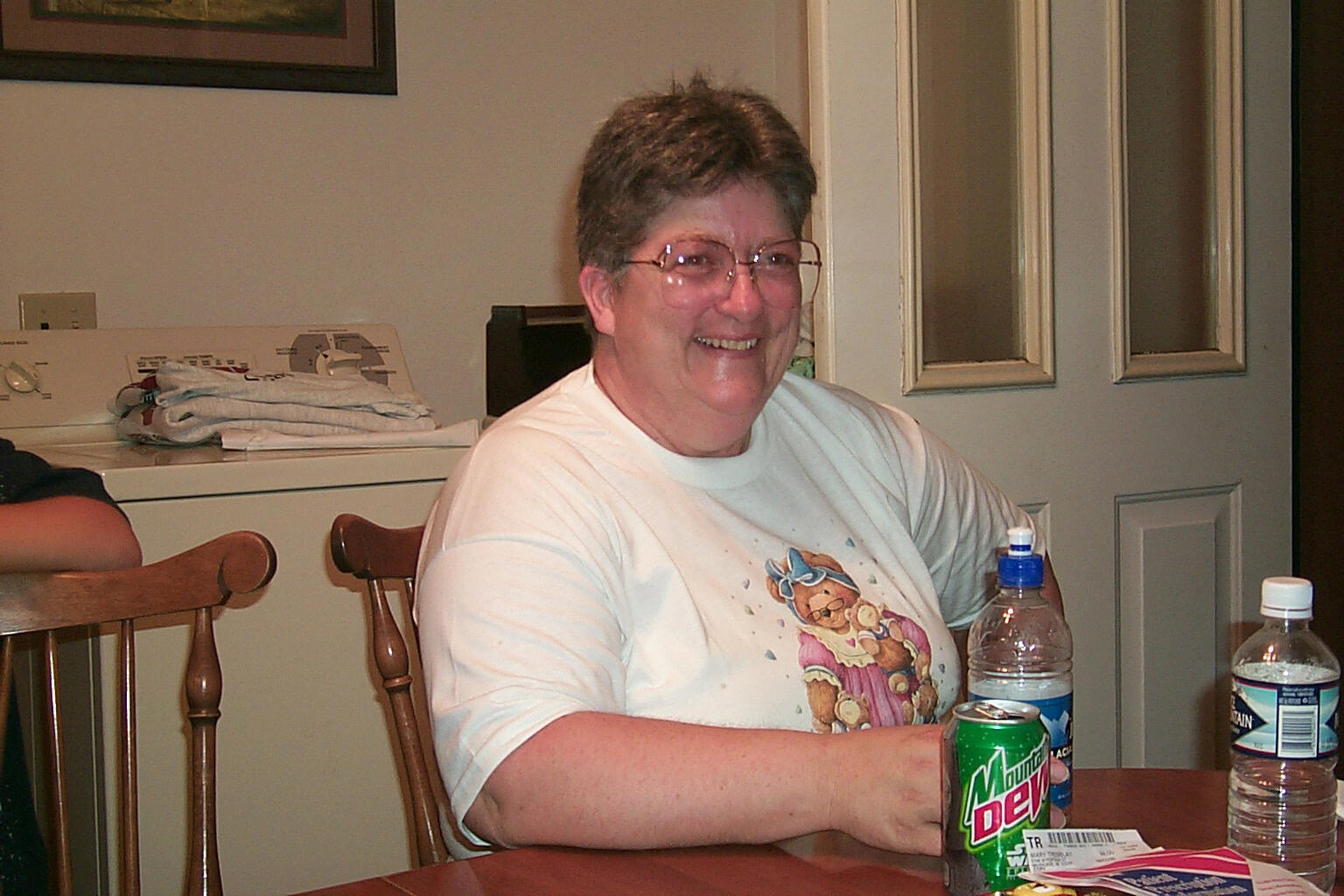 Smiling woman with short gray hair and glasses sits at a table with soda and water bottles nearby in a home setting.