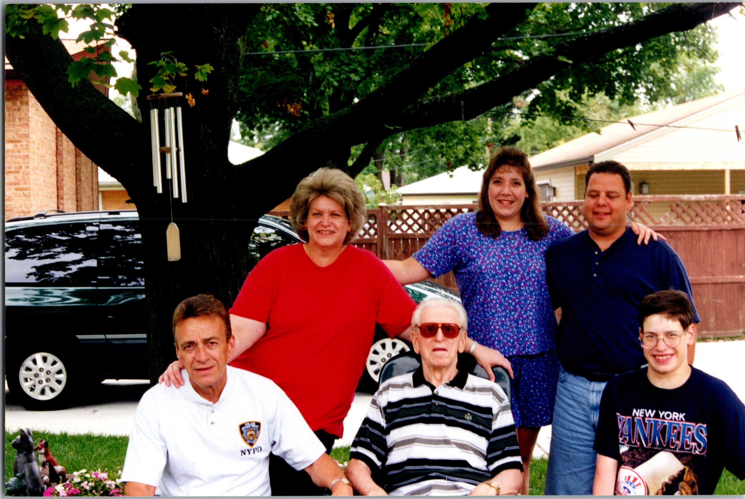 Group of people posing for a backyard photo under a large tree with wind chimes visible in the background.