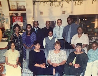 Diverse group of adults and children posing for a family-style group photo in a living room, seated on couches and chairs and smiling at the camera.