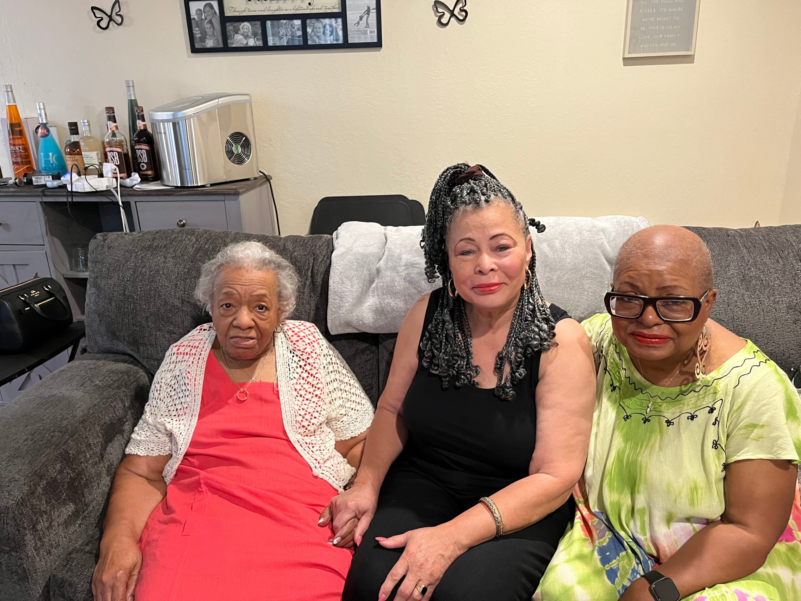 Three women sit closely on a gray sofa, smiling at the camera in a living room background with bottles and photos on a cabinet behind them.