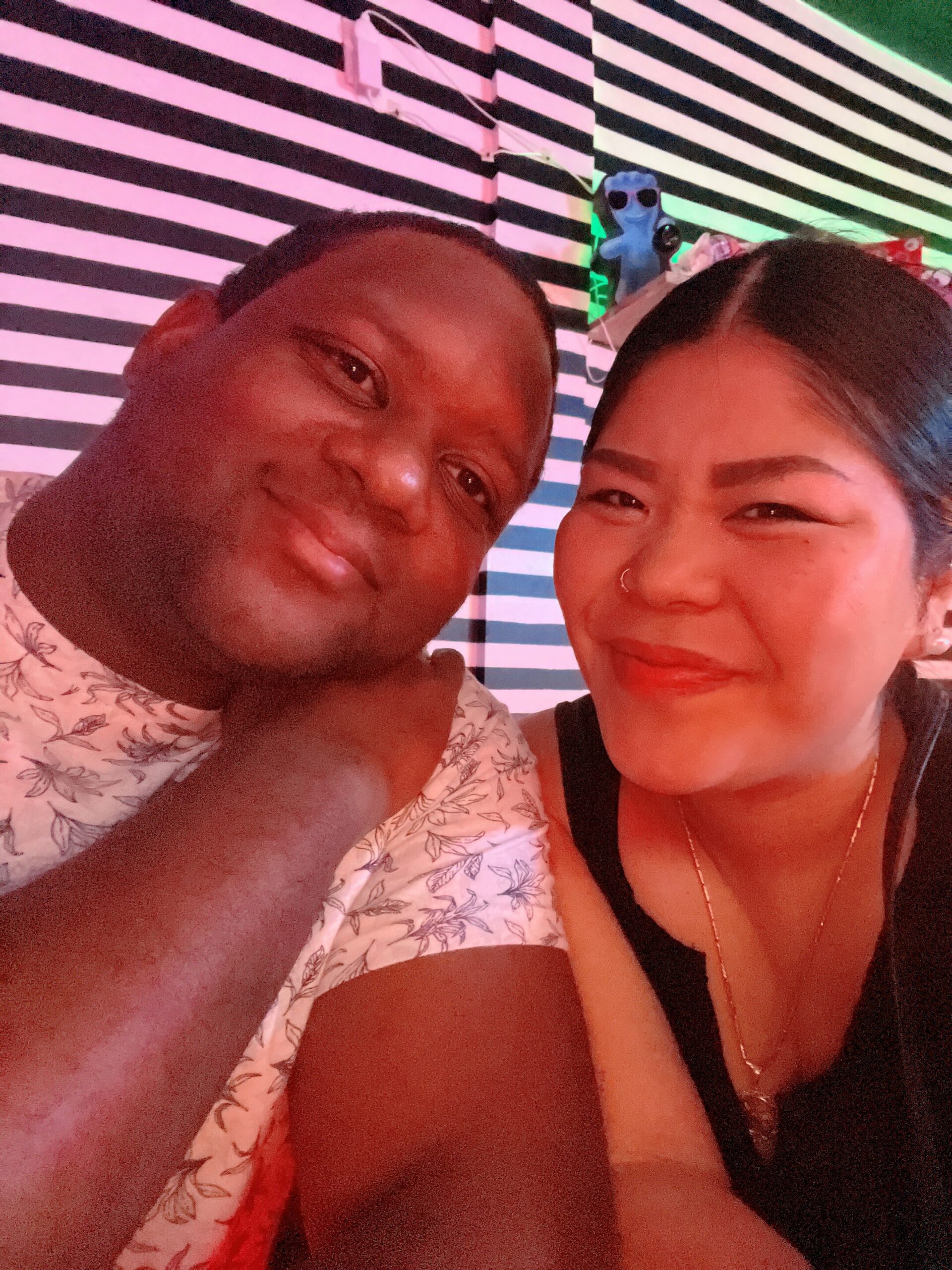 Two friends taking a close-up selfie under pink lighting, smiling at the camera with a striped wall behind them; man on the left in a floral shirt and a woman on the right with a nose ring and black top.