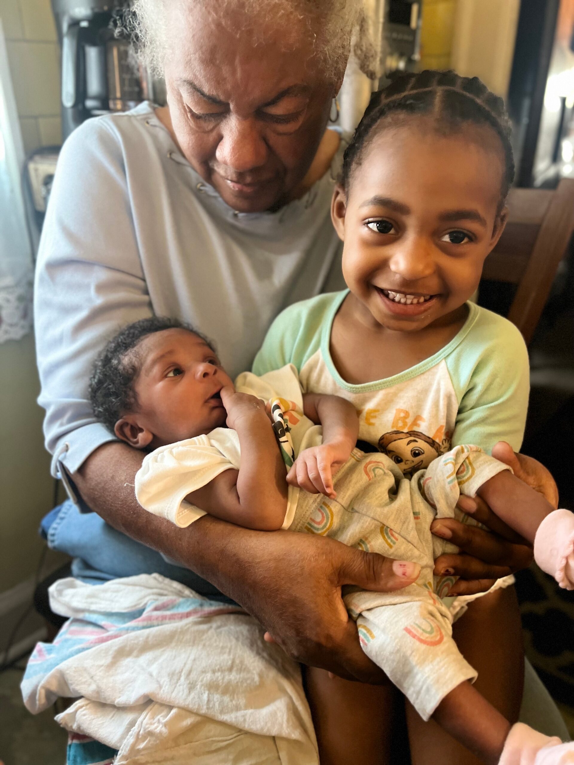 Elderly woman holding a newborn baby and a smiling toddler indoors.