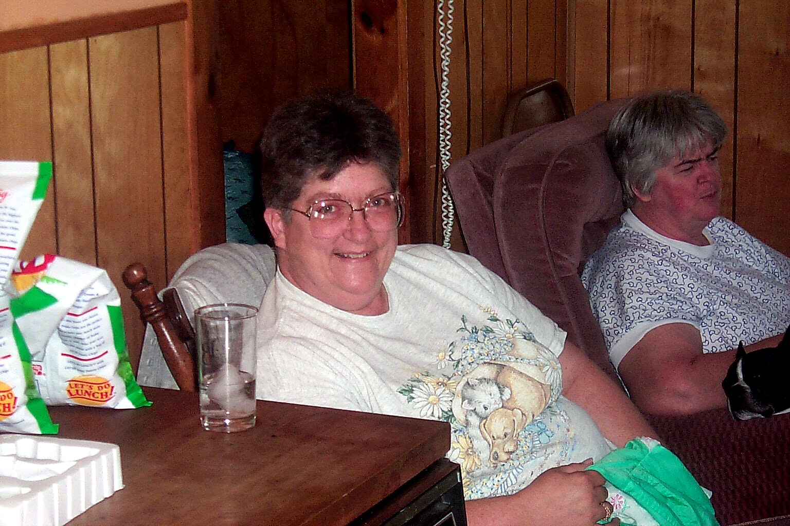 Smiling woman with short dark hair and glasses sits at a wooden table; a man reclines in a chair behind her in a wood-paneled room.