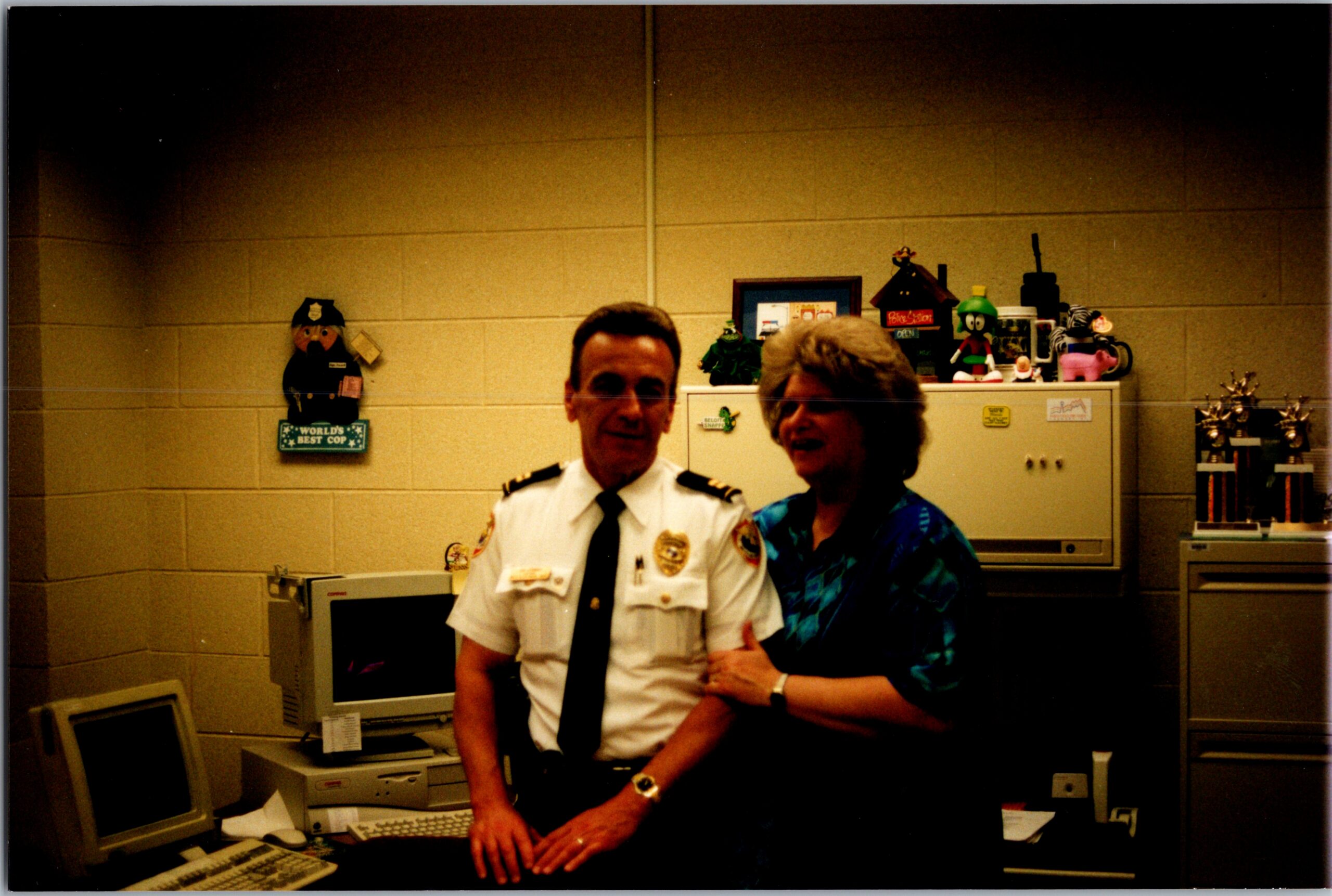 Police officer in uniform with a smiling woman, standing at a desk in an office with retro computer equipment and awards on the shelves behind them.