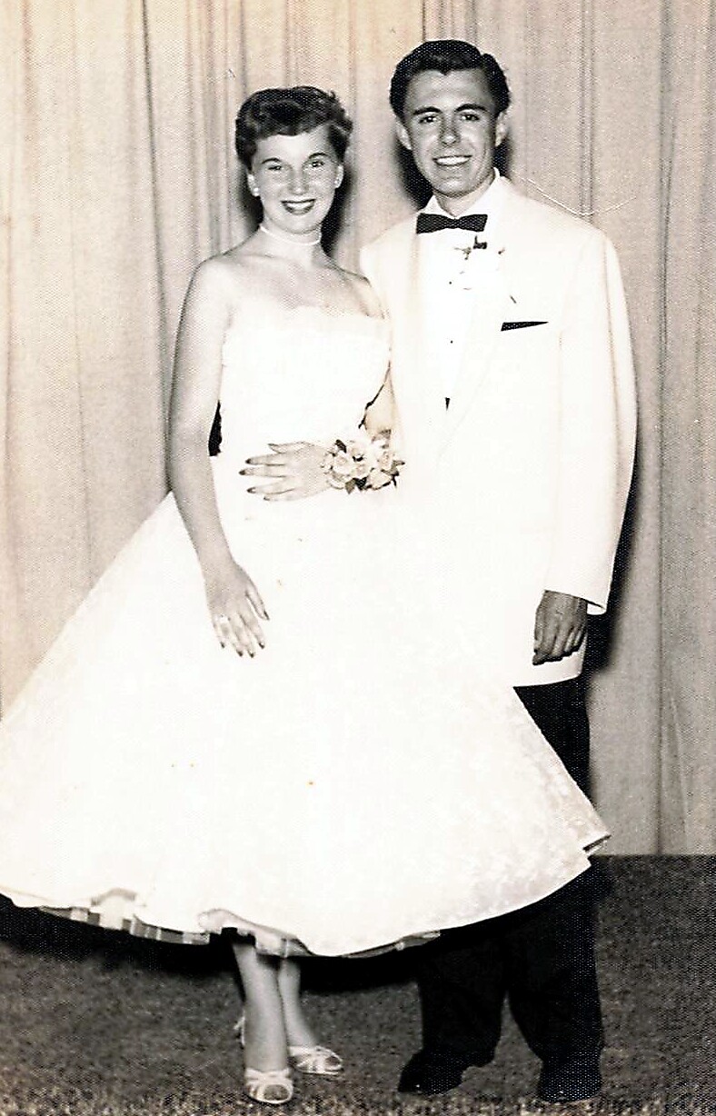 Black-and-white photo of a newlywed couple posing in formal wedding attire: bride in a strapless gown with bouquet, groom in a white tuxedo with bow tie.