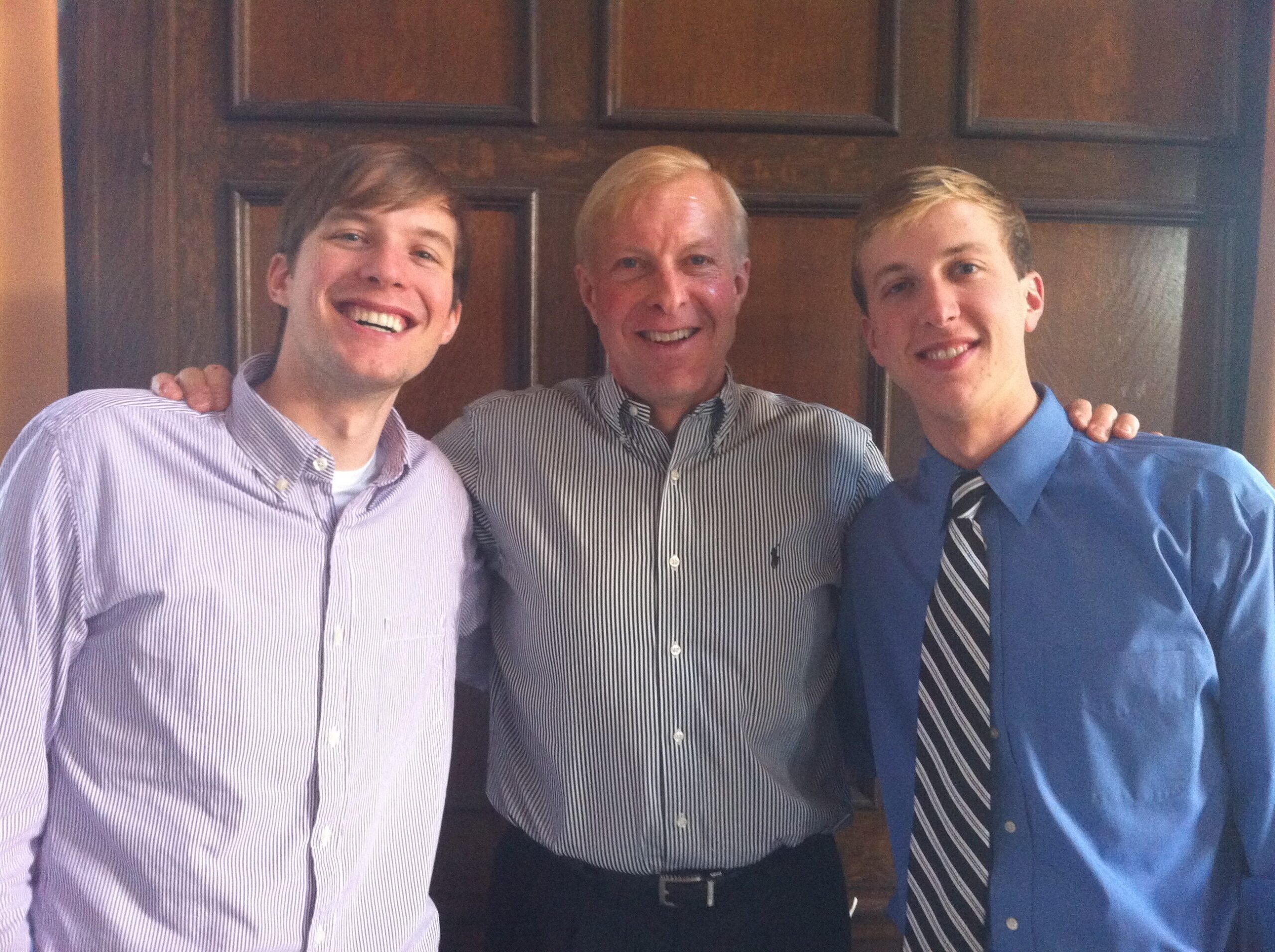 Three men in dress shirts smiling with arms around each other in front of a wooden door background.