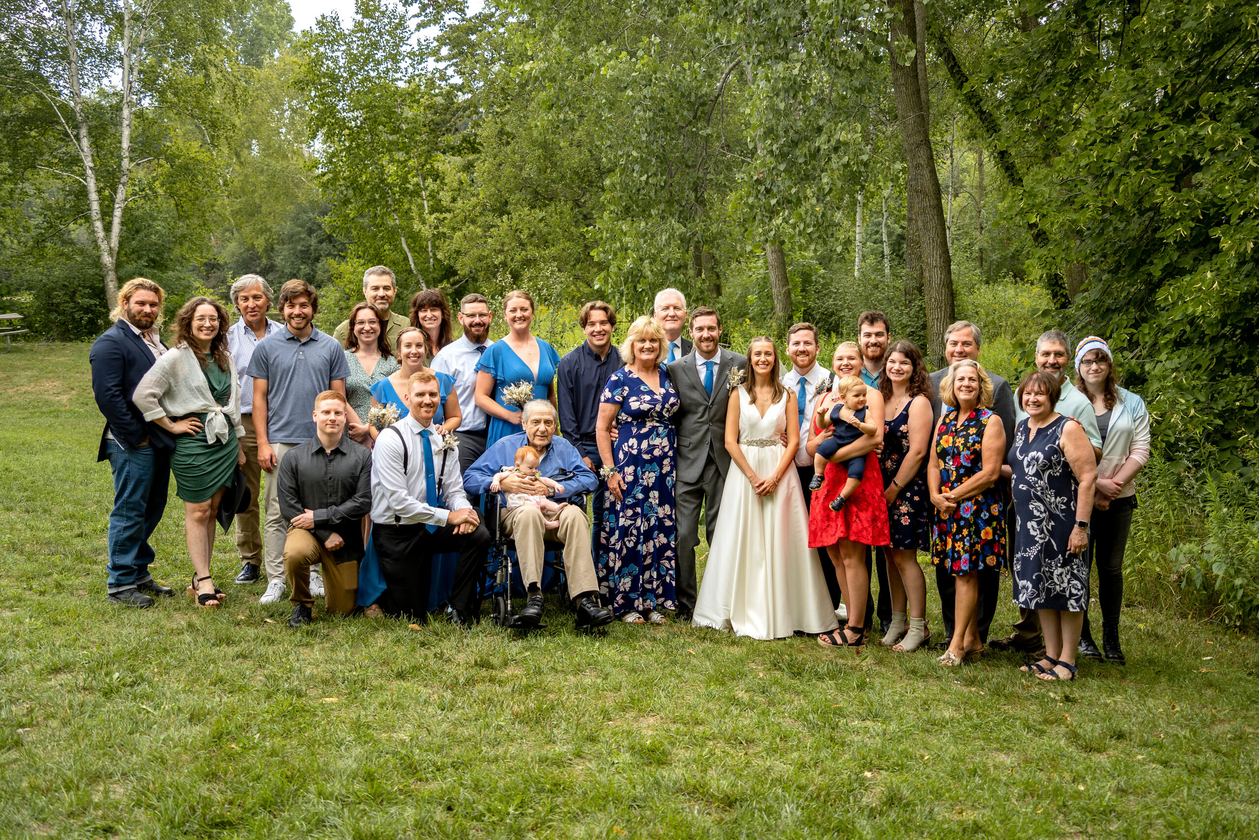 Large group portrait of wedding guests outdoors in a grassy park with trees in the background.