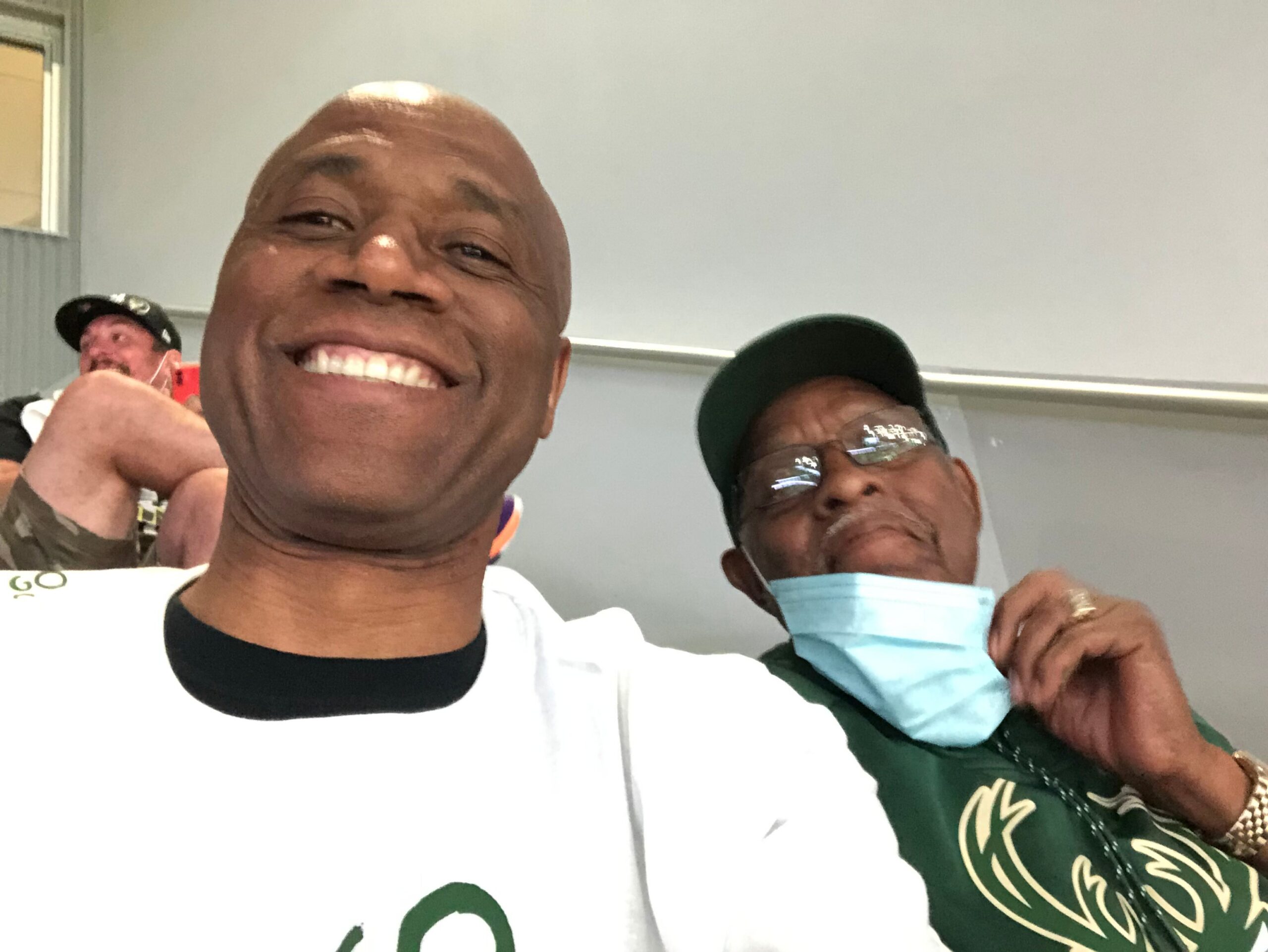 Two men taking a close-up selfie indoors; man in white shirt in foreground smiles widely, another man in a green shirt with a mask under his chin behind him.