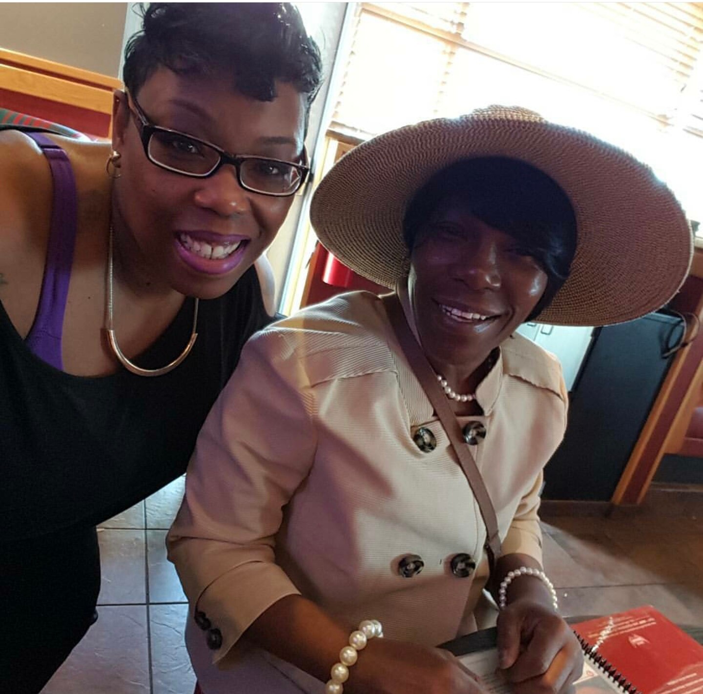 Two women sit side by side smiling for a selfie; the older woman wears a wide-brim beige hat and beige coat, jewelry visible.