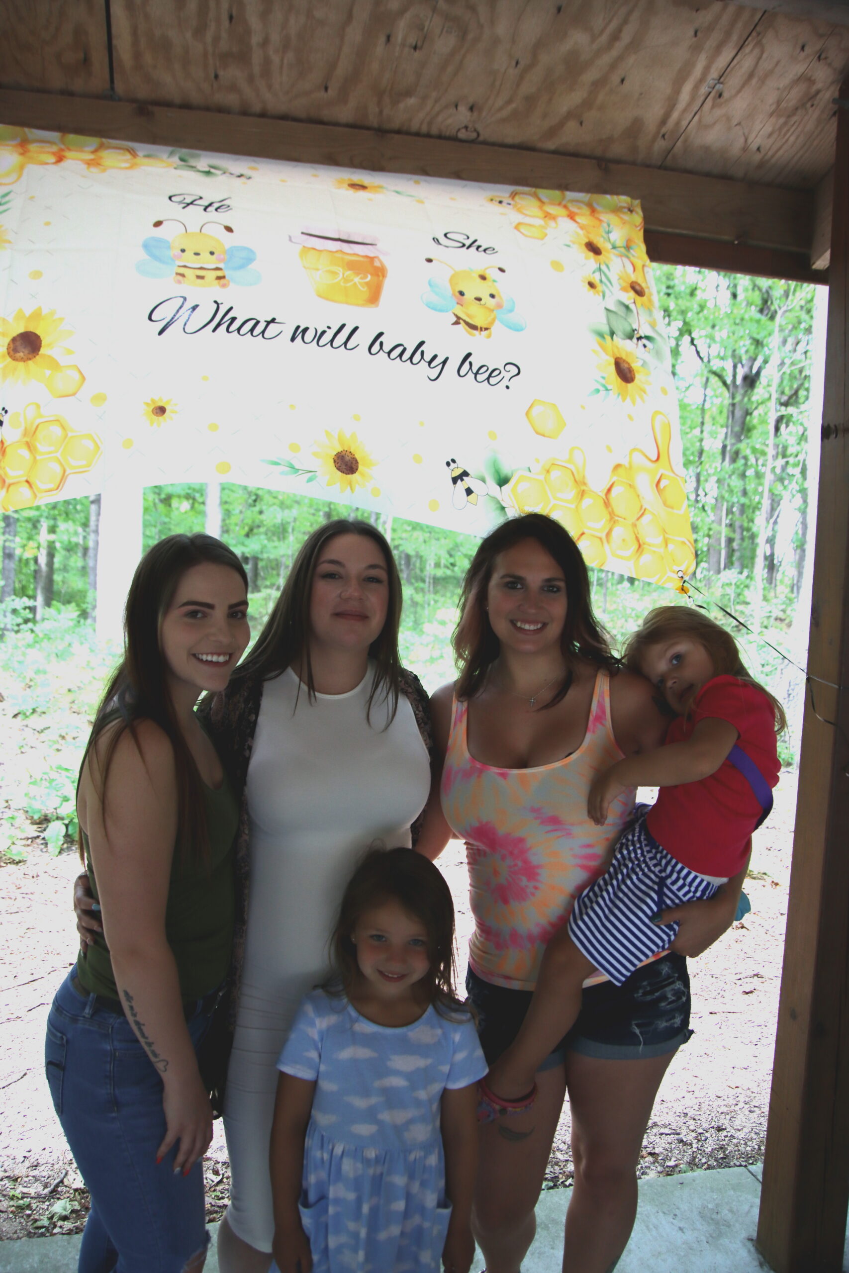 Group of women and children posing under a sunflower/bee-themed banner that reads 'What will baby bee?'