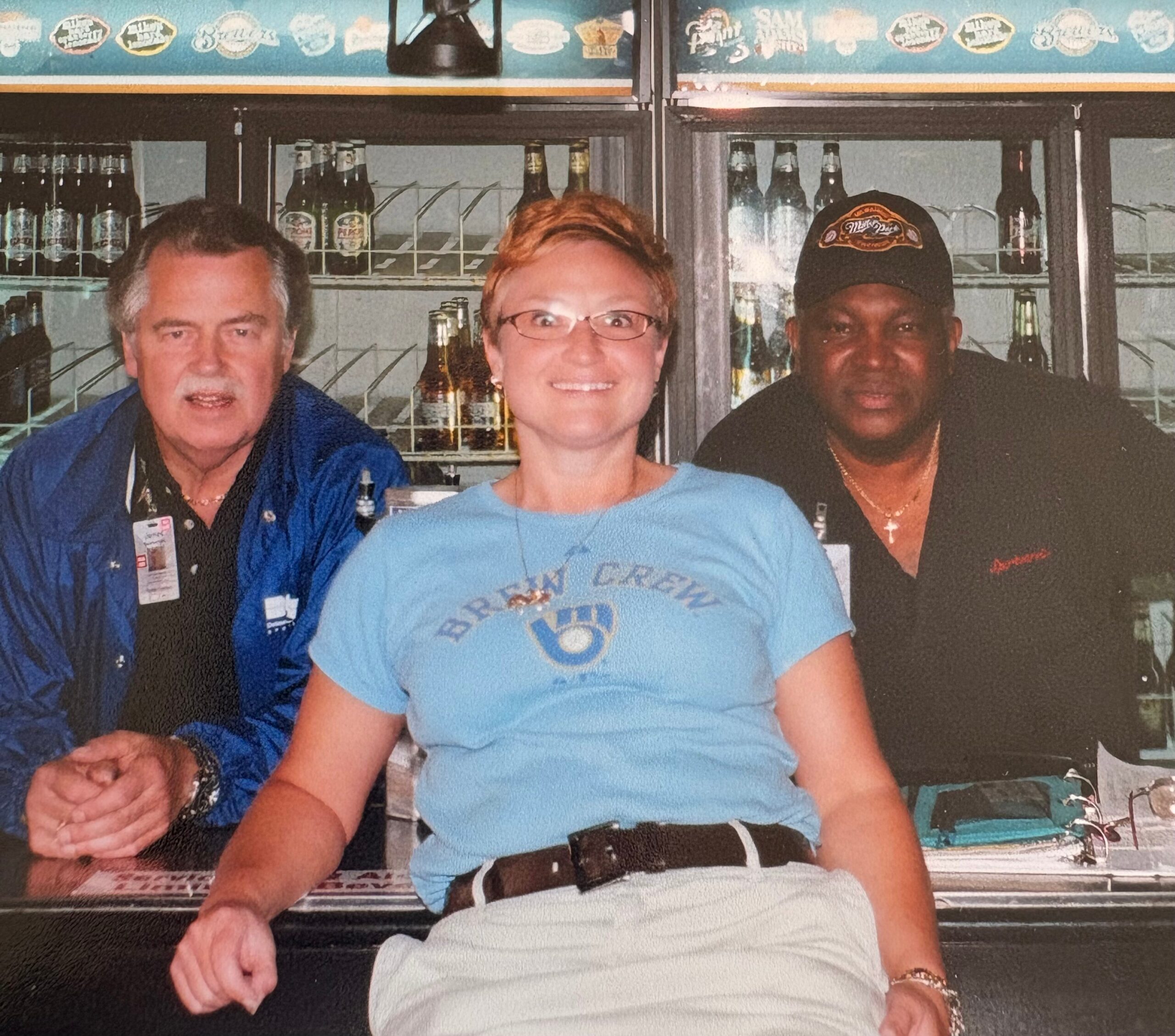 Three friends sit at a bar counter posing for a photo, with bottles on shelves behind them in a cooler.
