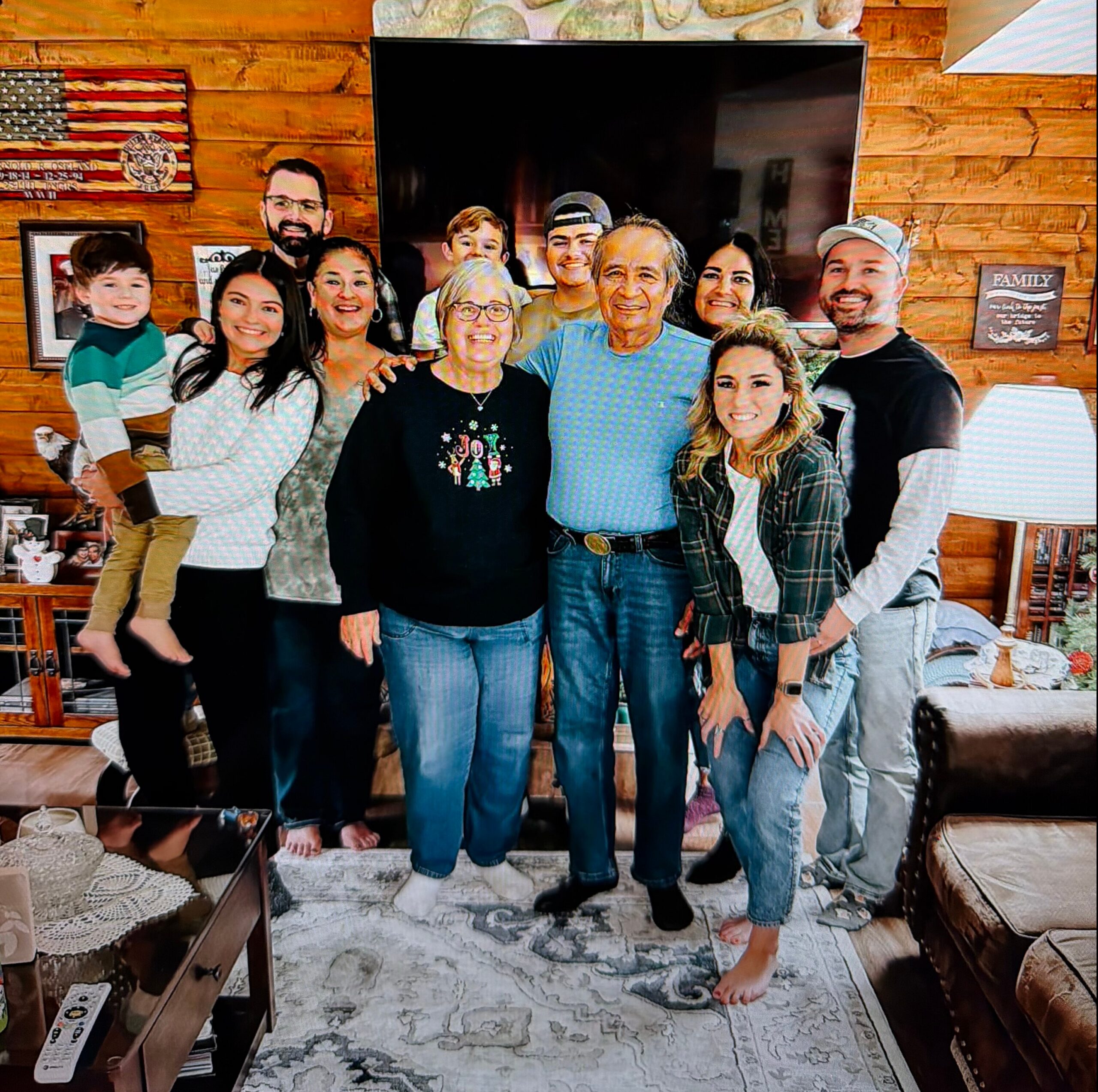 Group of about 12 friends and family posing together in a living room with a wooden wall and a large TV behind them, smiling at the camera.