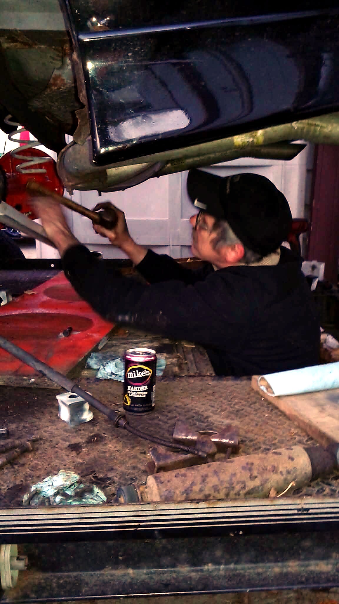 Mechanic under a raised car in a cluttered garage, using a tool while parts lie on the dusty floor.
