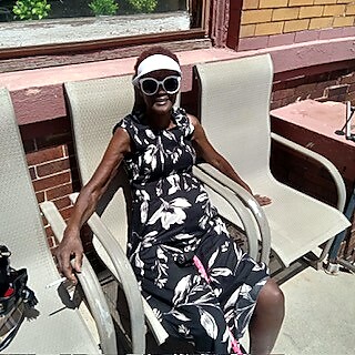 Older woman in a black-and-white floral dress, white visor, and sunglasses sitting in a patio lounge chair outdoors.