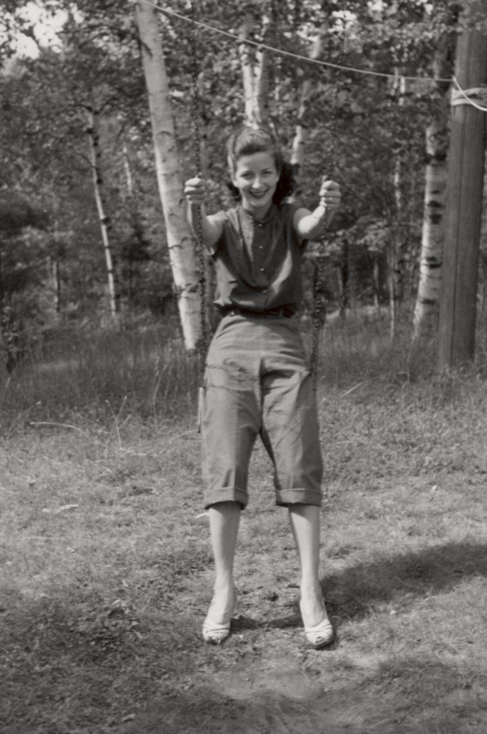 Girl on a swing smiling at the camera in a wooded yard with trees behind her.