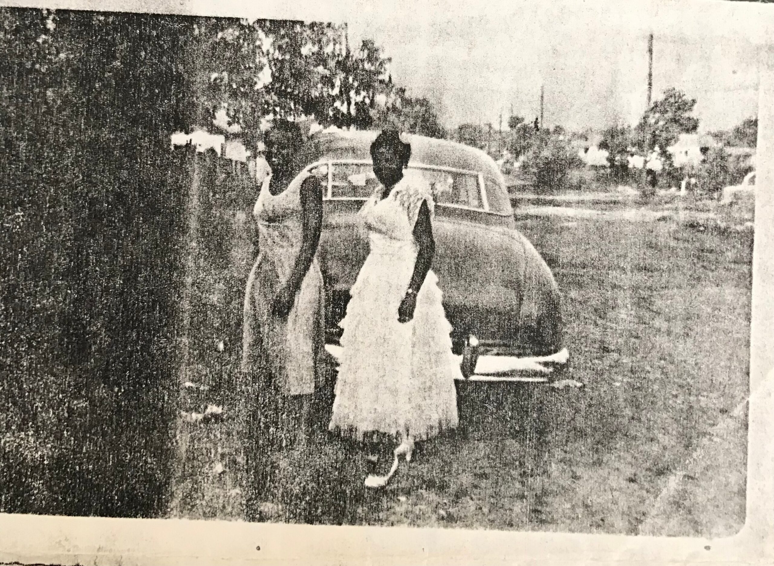 Two people pose beside a vintage car in an open, grassy area (black-and-white photo).
