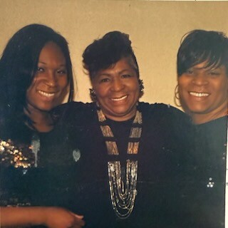 Three smiling women posing together for a photo indoors, wearing dark outfits; left in a sequined top, middle with a layered necklace, right with short hair and hoop earrings.