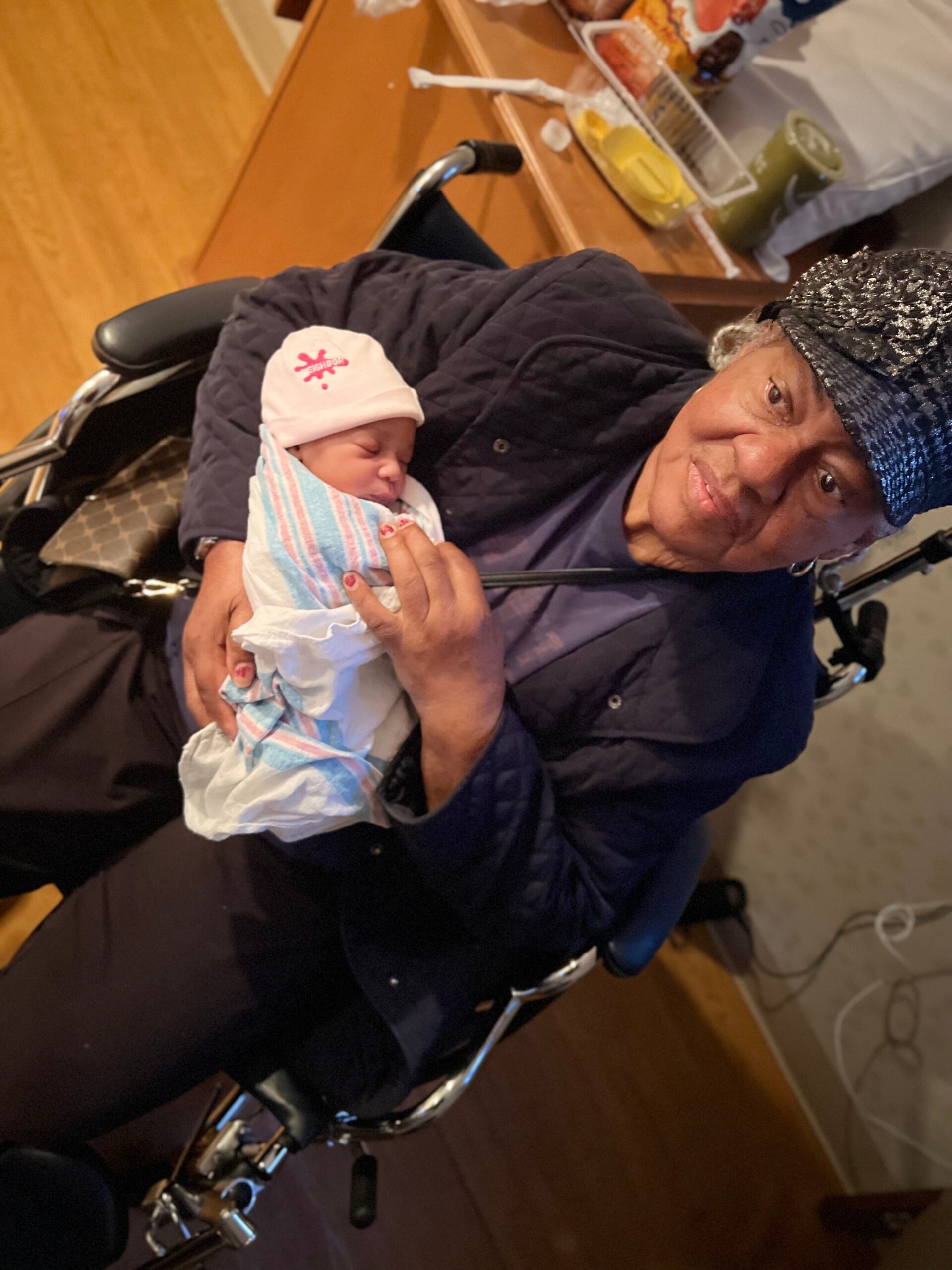 Older person in a wheelchair gently holds a newborn baby wrapped in a blanket.