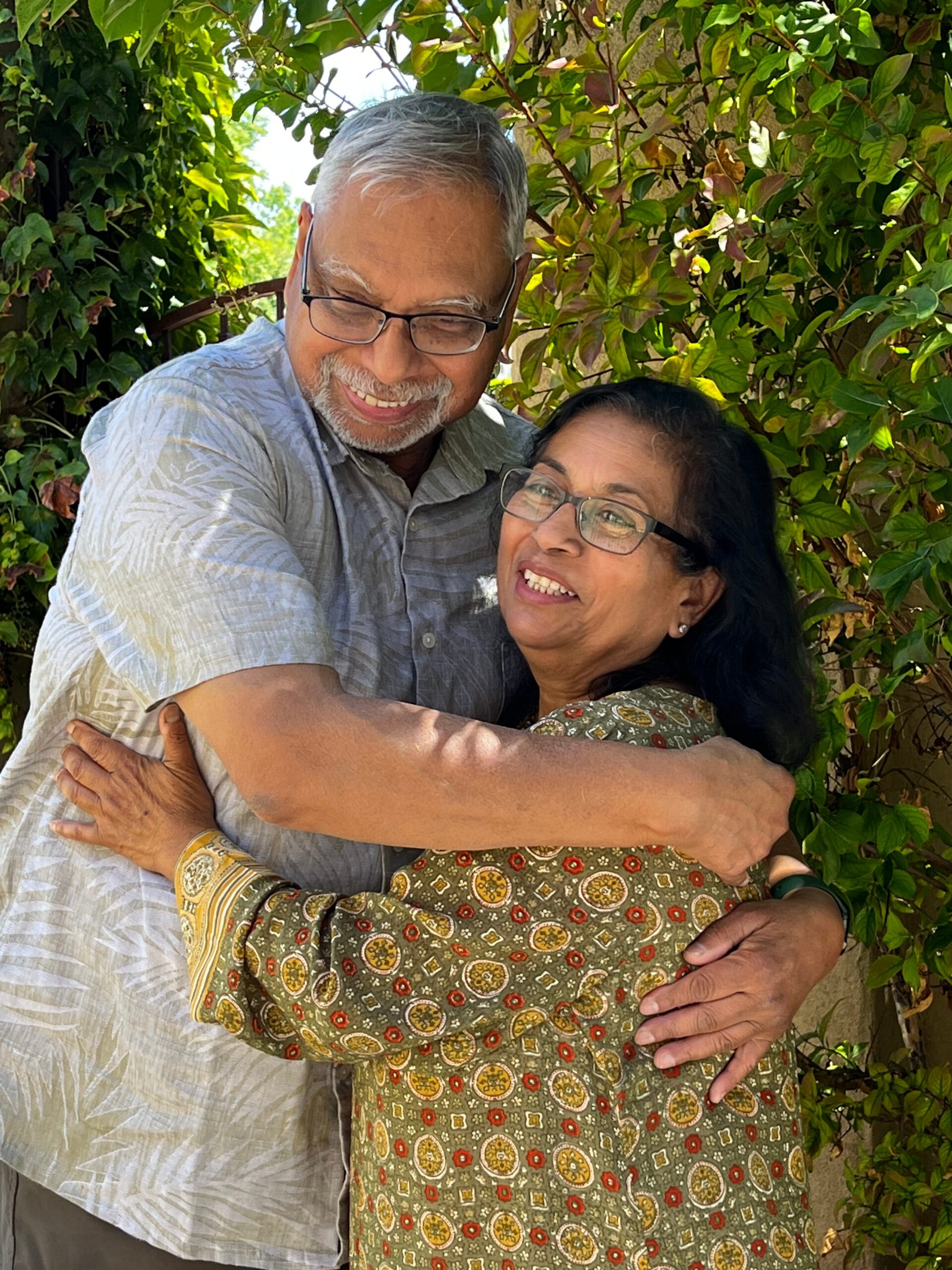 An older couple smiling and embracing outdoors among green leafy bushes.