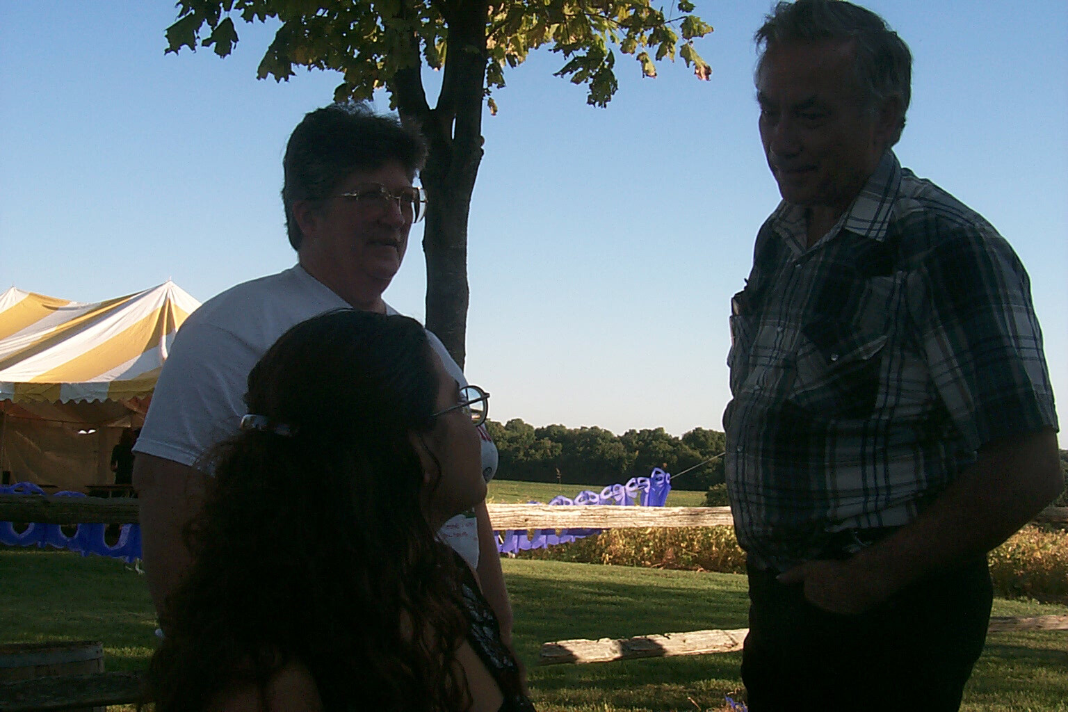 Three people converse outdoors in a park near a striped yellow tent and blue chairs under a tree.