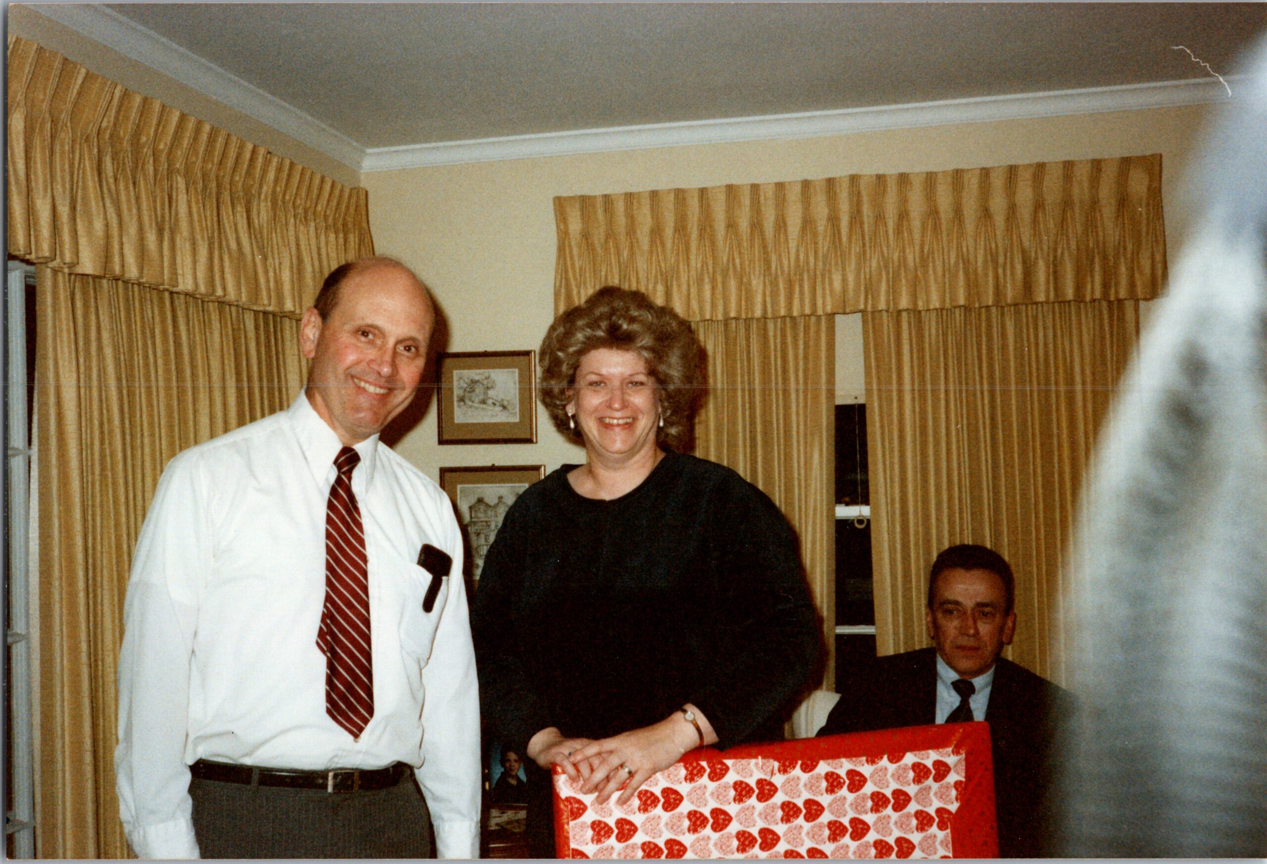 Man in a white shirt and striped tie stands beside a smiling woman in a black dress, with a heart-patterned gift box in the foreground and gold curtains in the background.