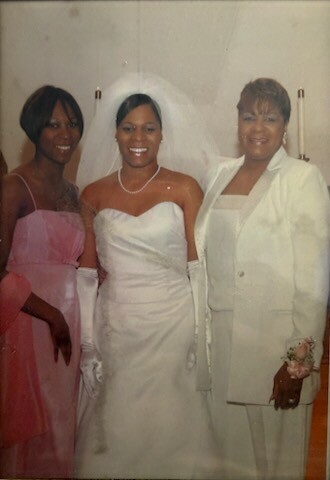 Three women posing together at a wedding: the center woman is the bride in a white gown and veil, flanked by two guests in formal dresses (one pink, one white).