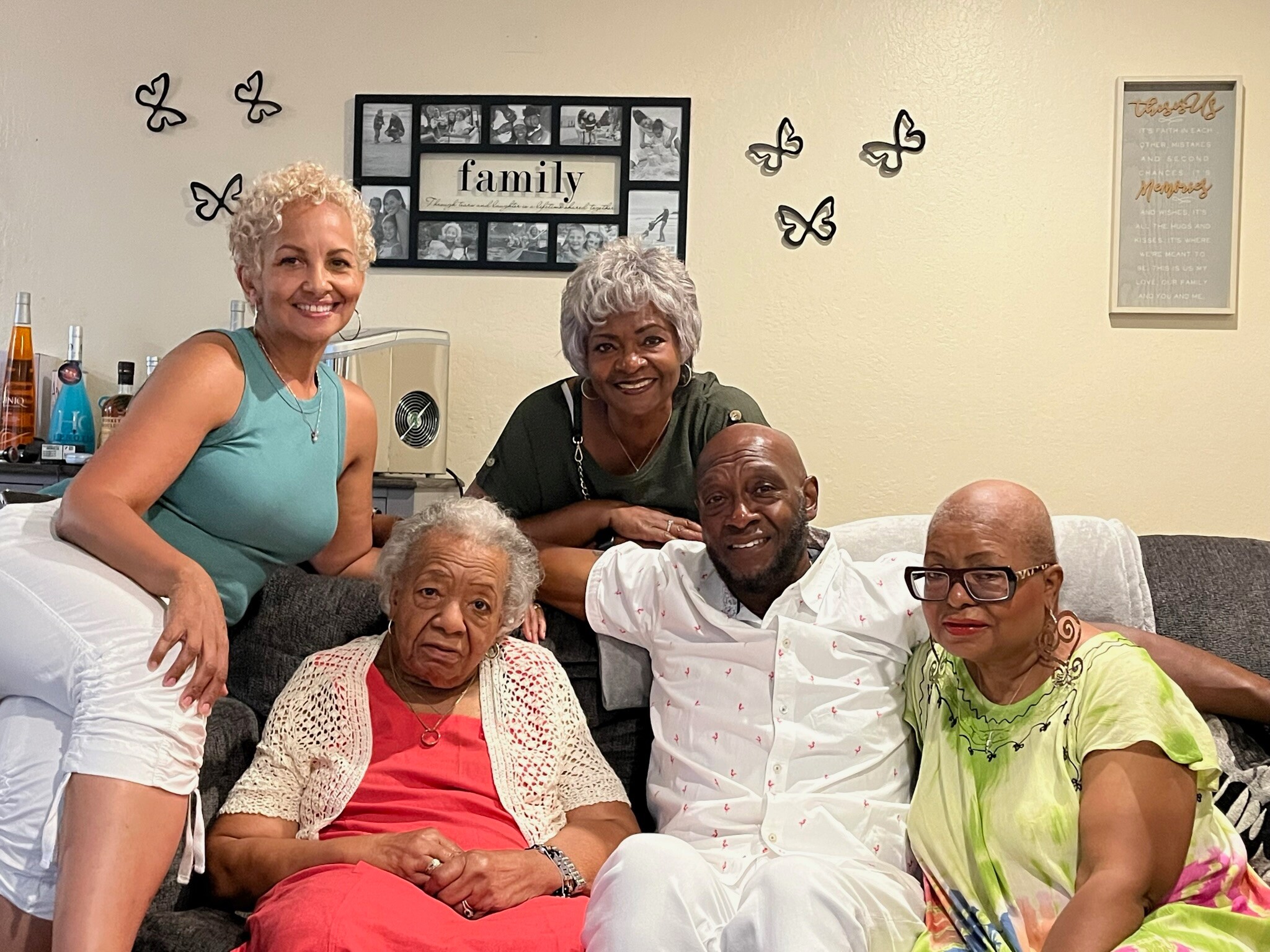 Six adults gathered on a living-room couch, smiling for the camera with butterfly wall decor in the background.
