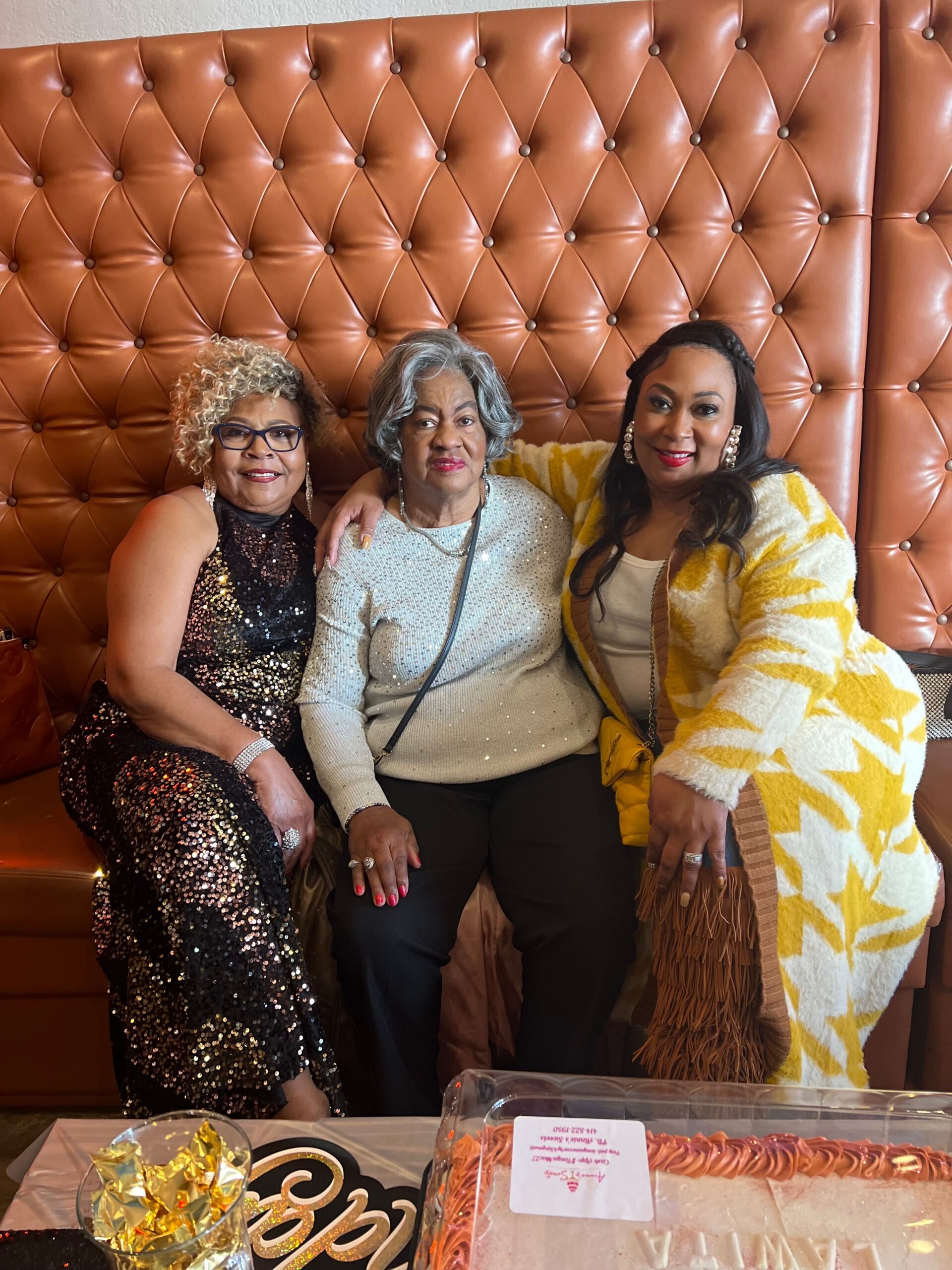 Three women pose together on a tufted brown leather bench at a celebration, with a cake and snacks in the foreground.