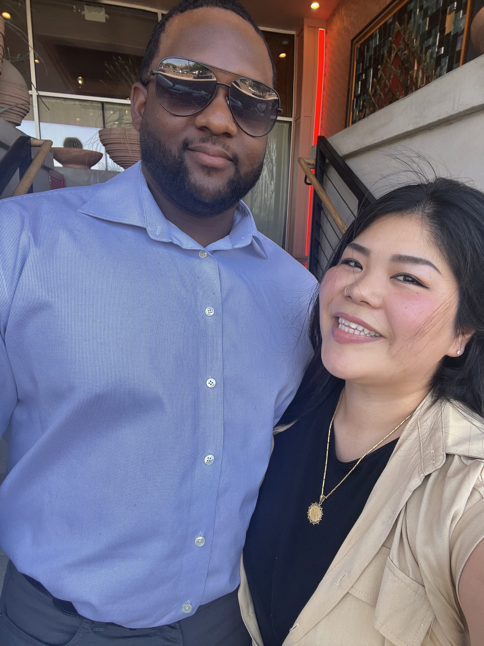 A smiling man in a light blue button-up shirt and sunglasses stands beside a smiling woman in a beige blazer and black top wearing a gold pendant; they pose for a selfie outdoors.