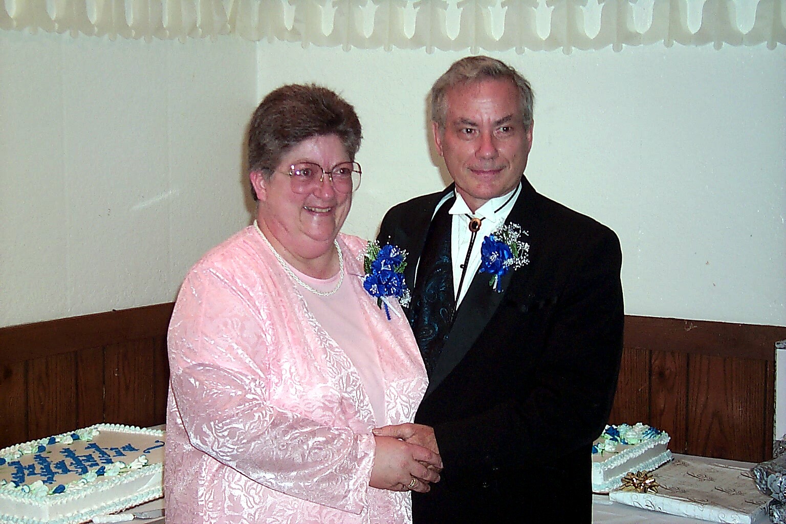 Smiling older couple in formal attire posing together at a celebration, with a decorated cake in the background.
