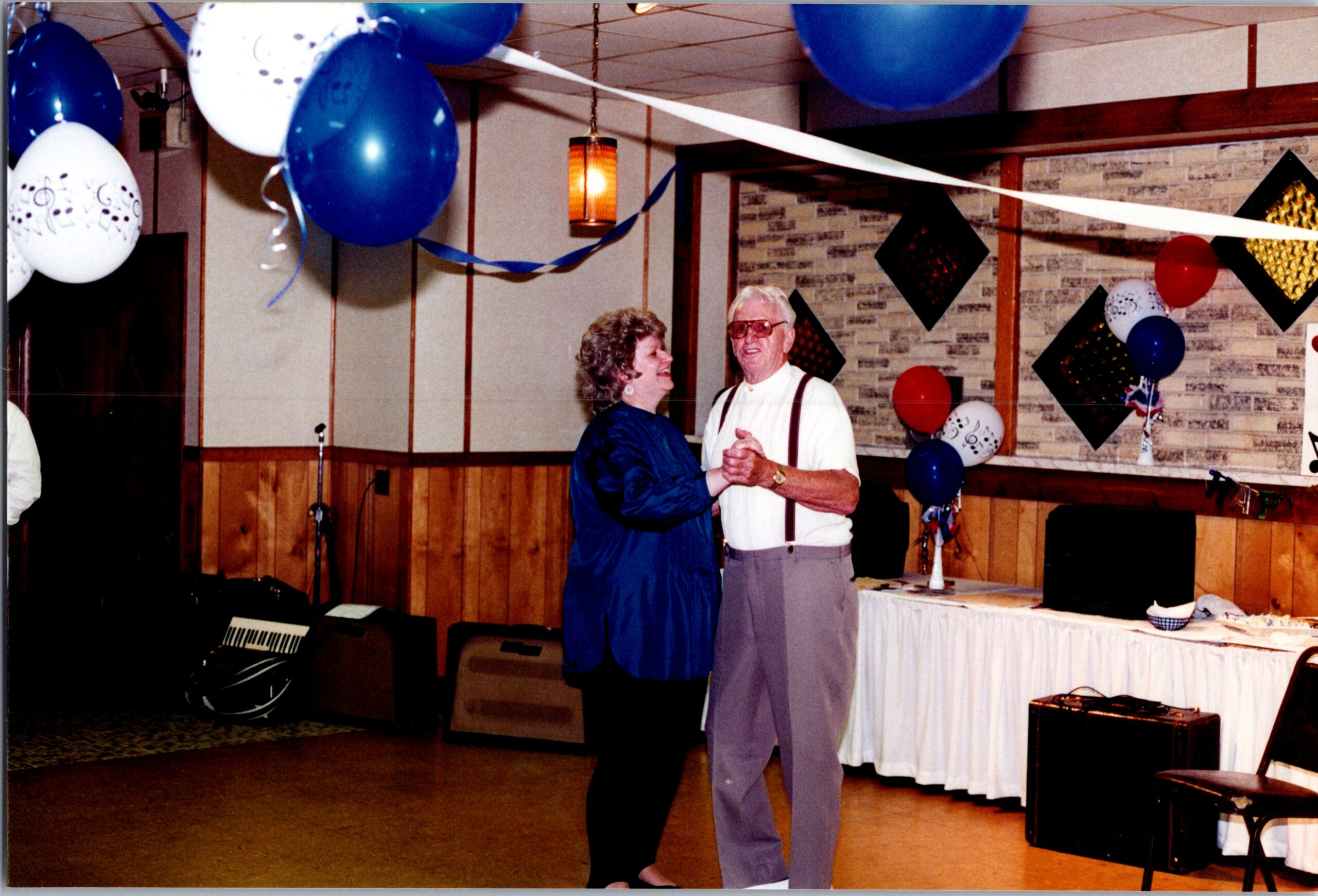 An elderly couple dances together at a party, surrounded by blue and white balloons and streamers in a decorated hall