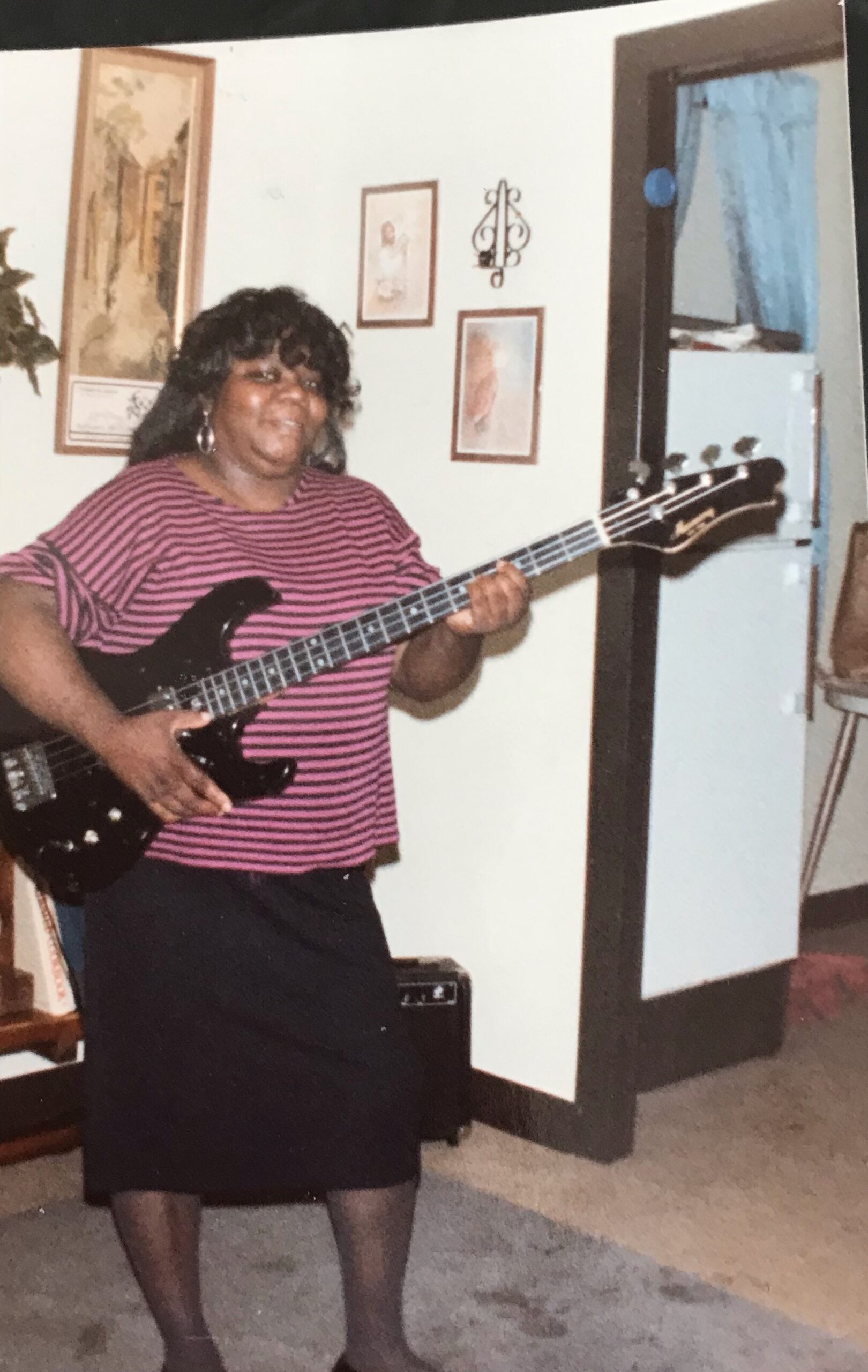 Woman with curly dark hair wearing a pink and black striped shirt playing a black bass guitar in a living room.
