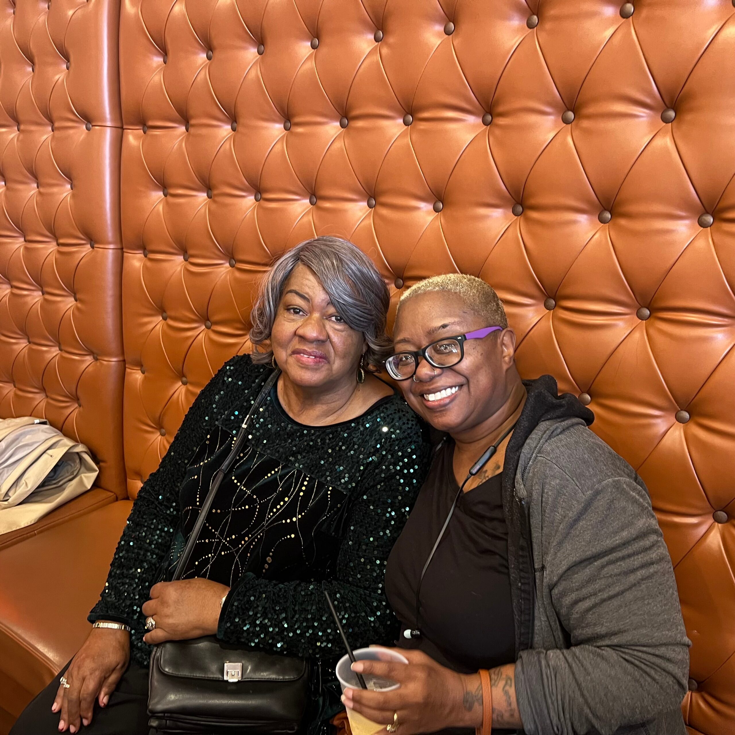 Two smiling women sit close together in a tufted orange booth, one in a black sequined top and the other wearing glasses and a gray hoodie.