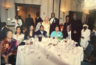 Group of about 14 people in formal attire seated around a round, white-tablecloth banquet table, posing for a photo in a dining hall.