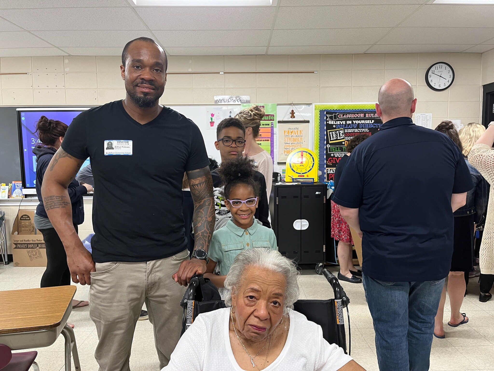 A classroom scene with a man in a black shirt and a girl in a wheelchair at the foreground, and several adults in the background near bulletin boards and a clock on the wall.
