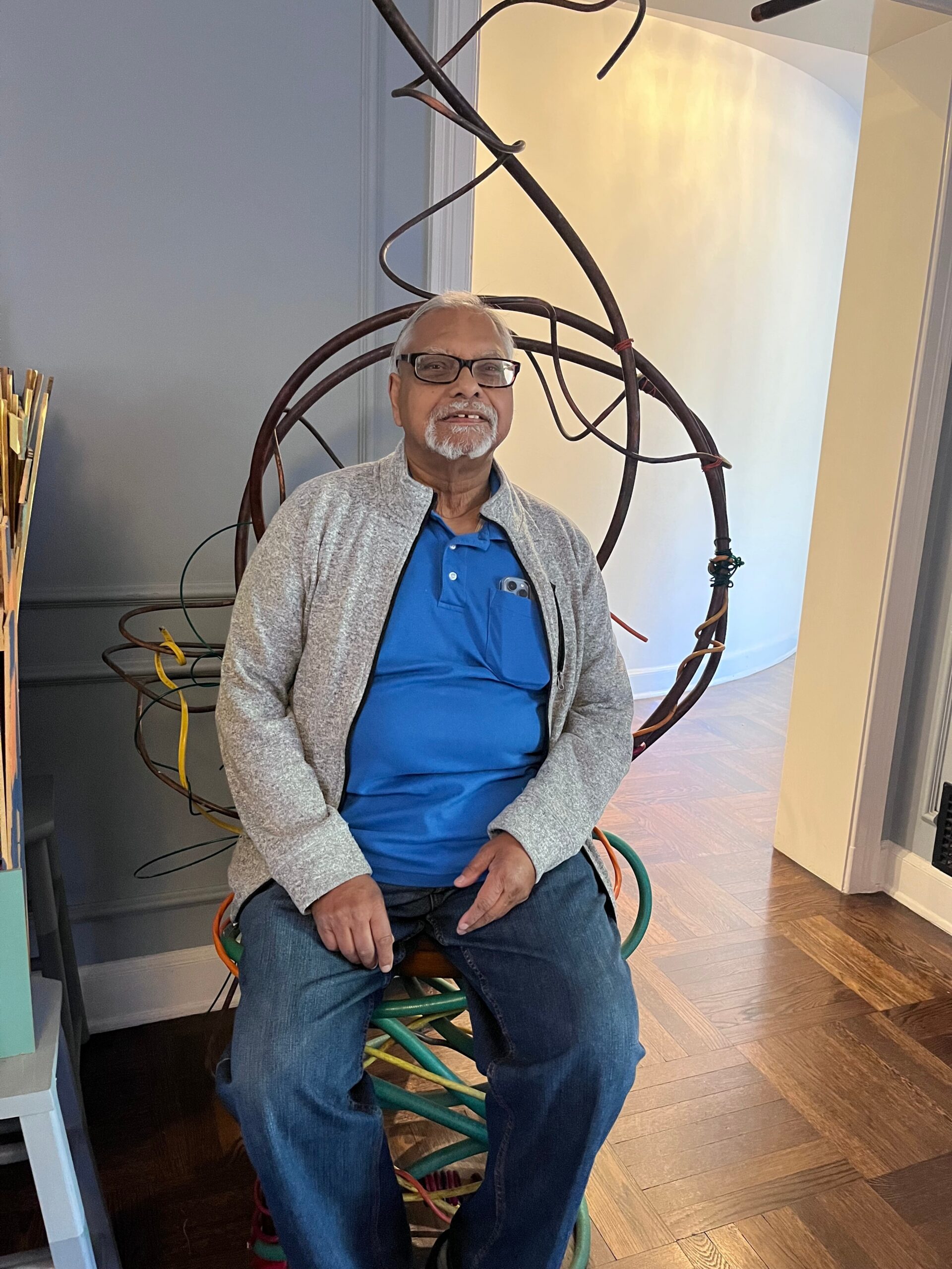 Older man with glasses and gray beard sits in front of a tangled metal wire sculpture indoors.