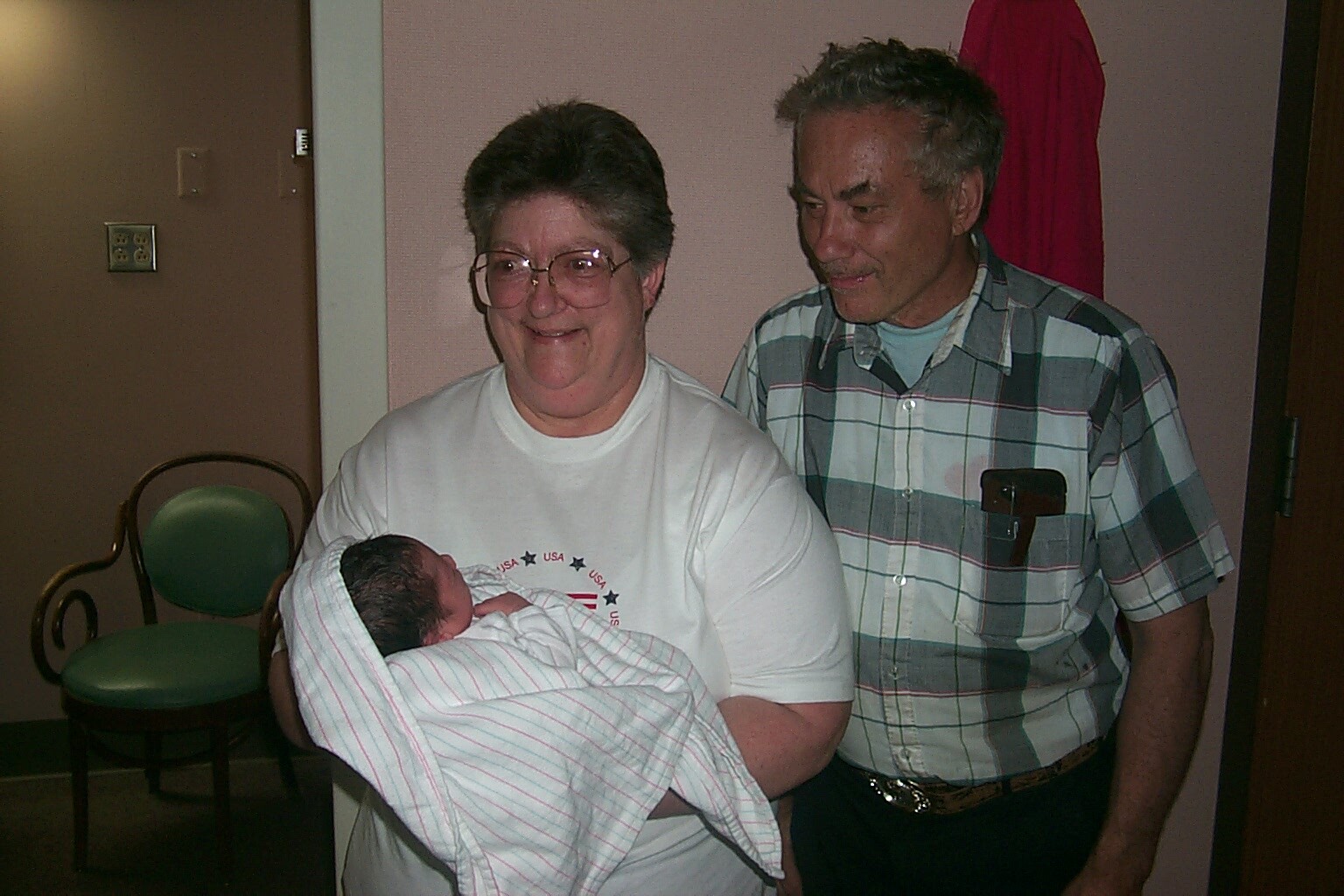 Smiling grandmother holds a newborn wrapped in a striped blanket as grandfather stands beside her in a doorway.