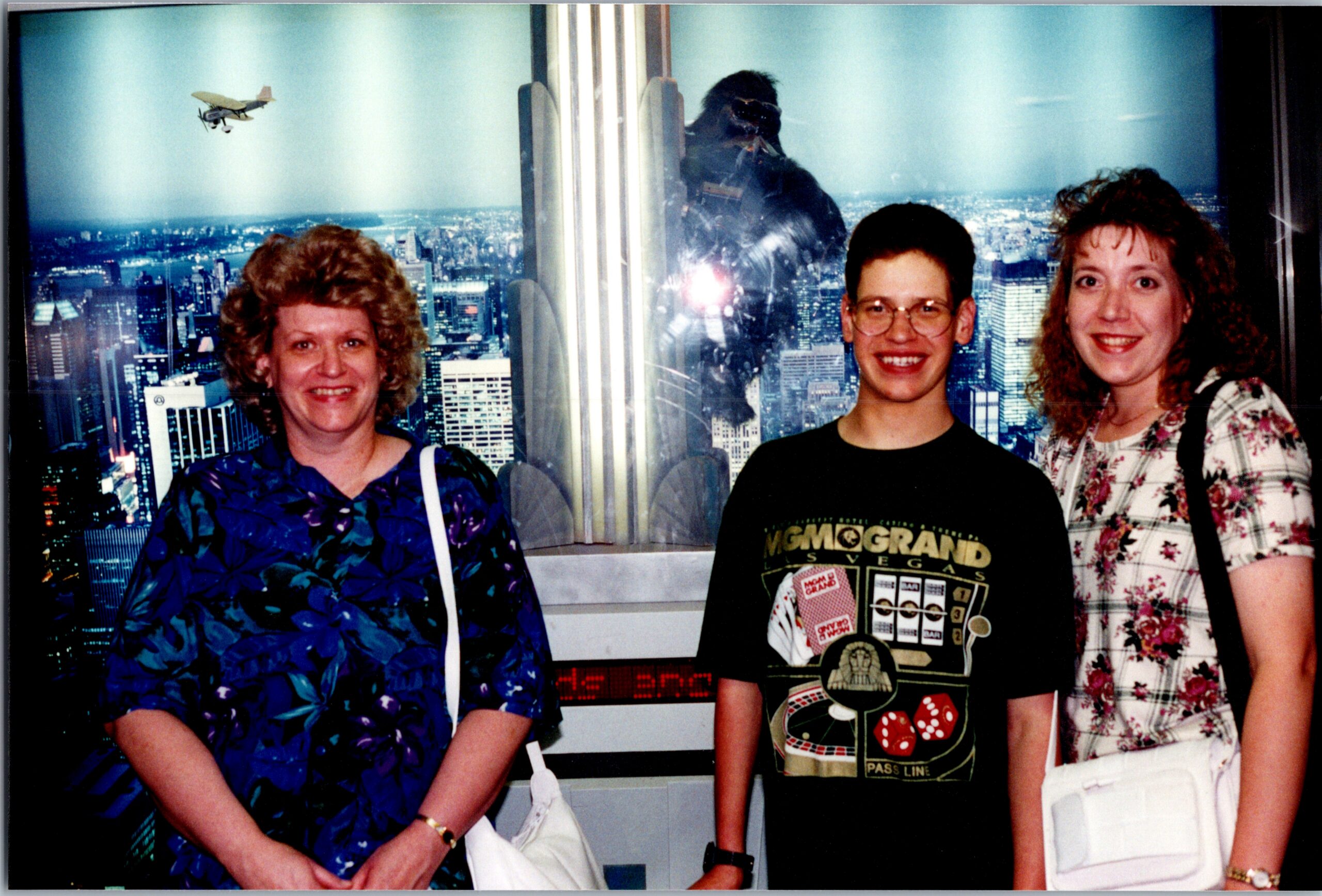 Three smiling women pose together indoors with a city skyline backdrop behind them.